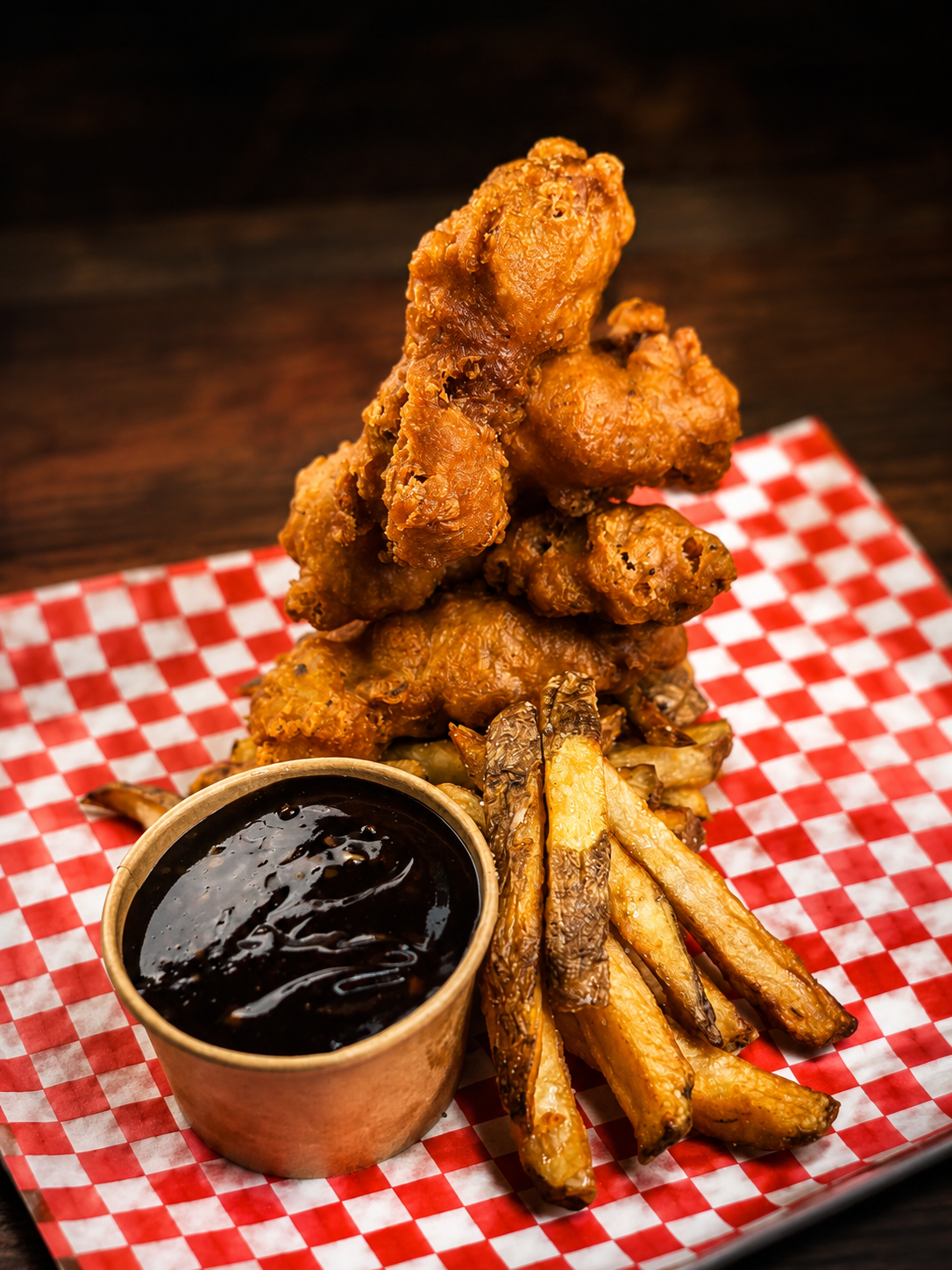 Fried chicken wings arranged in a tower on a red checkered paper with a side of French fries and a small cup of dipping sauce.