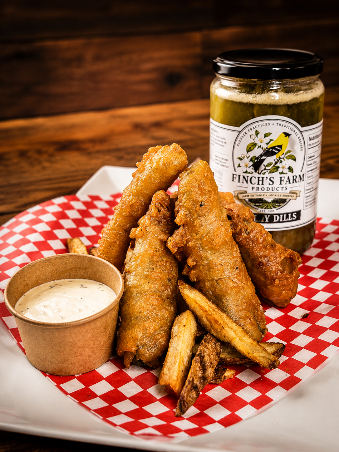 Fried chicken wings with fries served with dipping sauce, pickle relish, and a jar of pickles on a checkered paper in a white serving tray.
