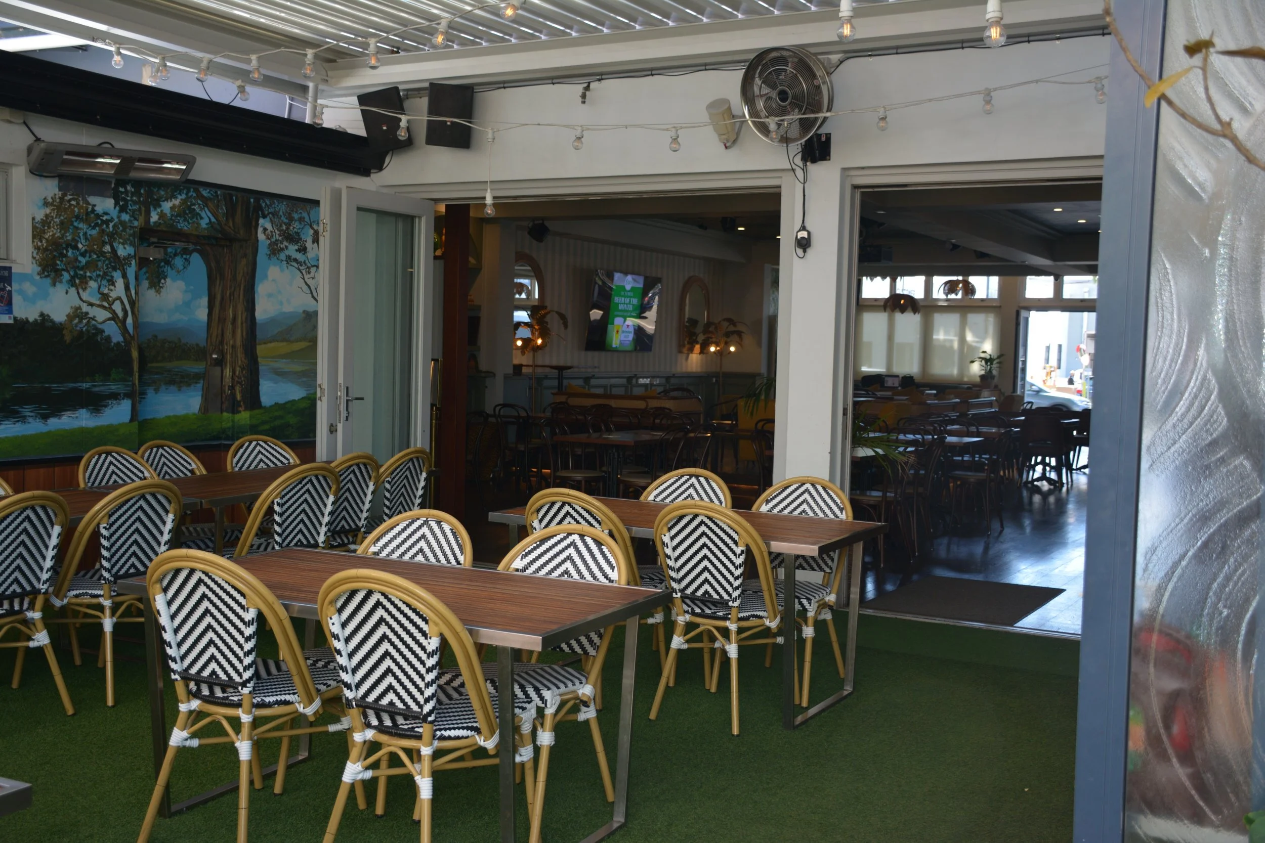 Empty indoor restaurant seating area with tables and chairs, a mural of a tree by a lake, and a large window showing outside, decorated with string lights and plants.