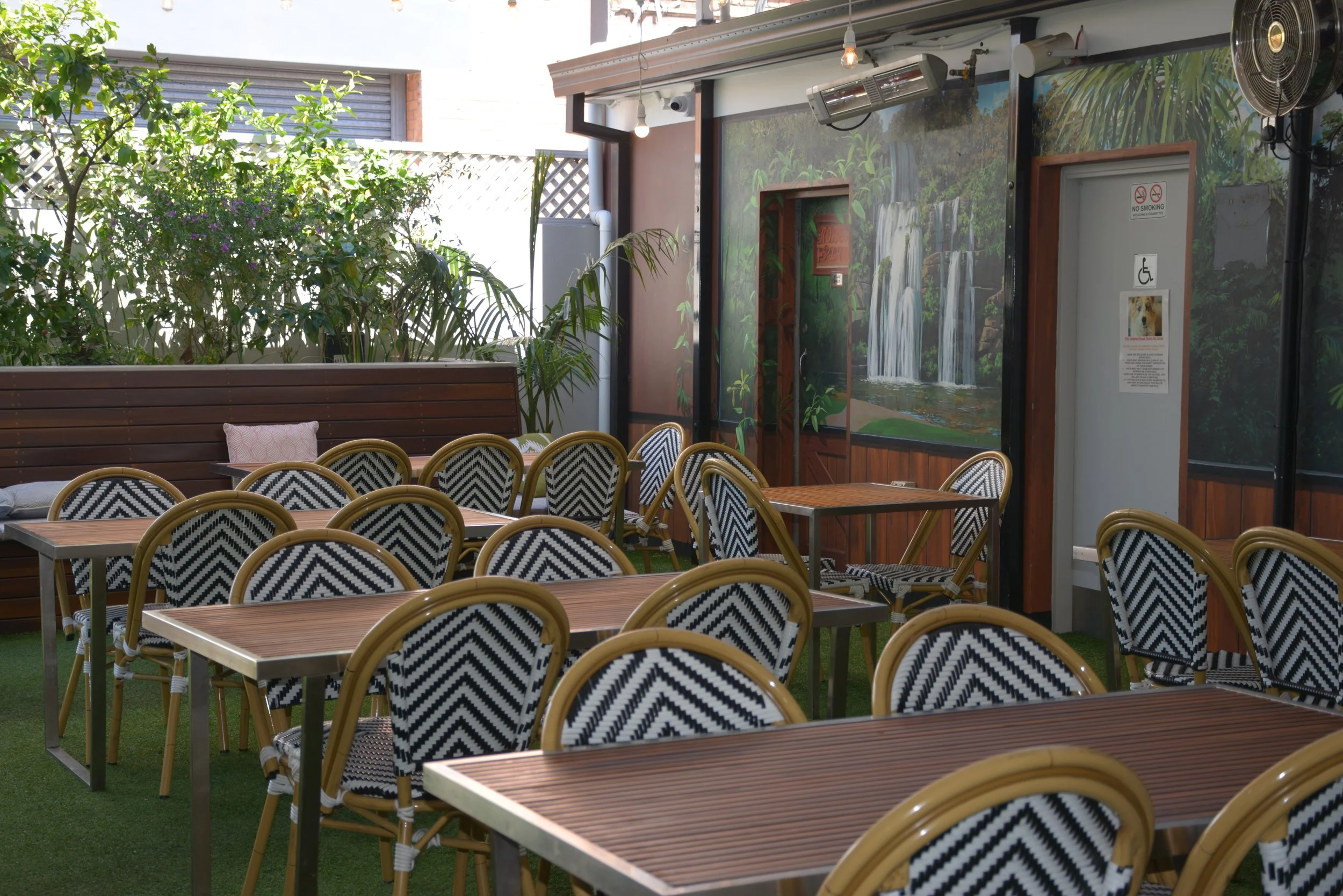 Outdoor patio with wooden tables and chairs with black and white zigzag patterned cushions, surrounded by greenery and plants, and a wall with a waterfall mural.