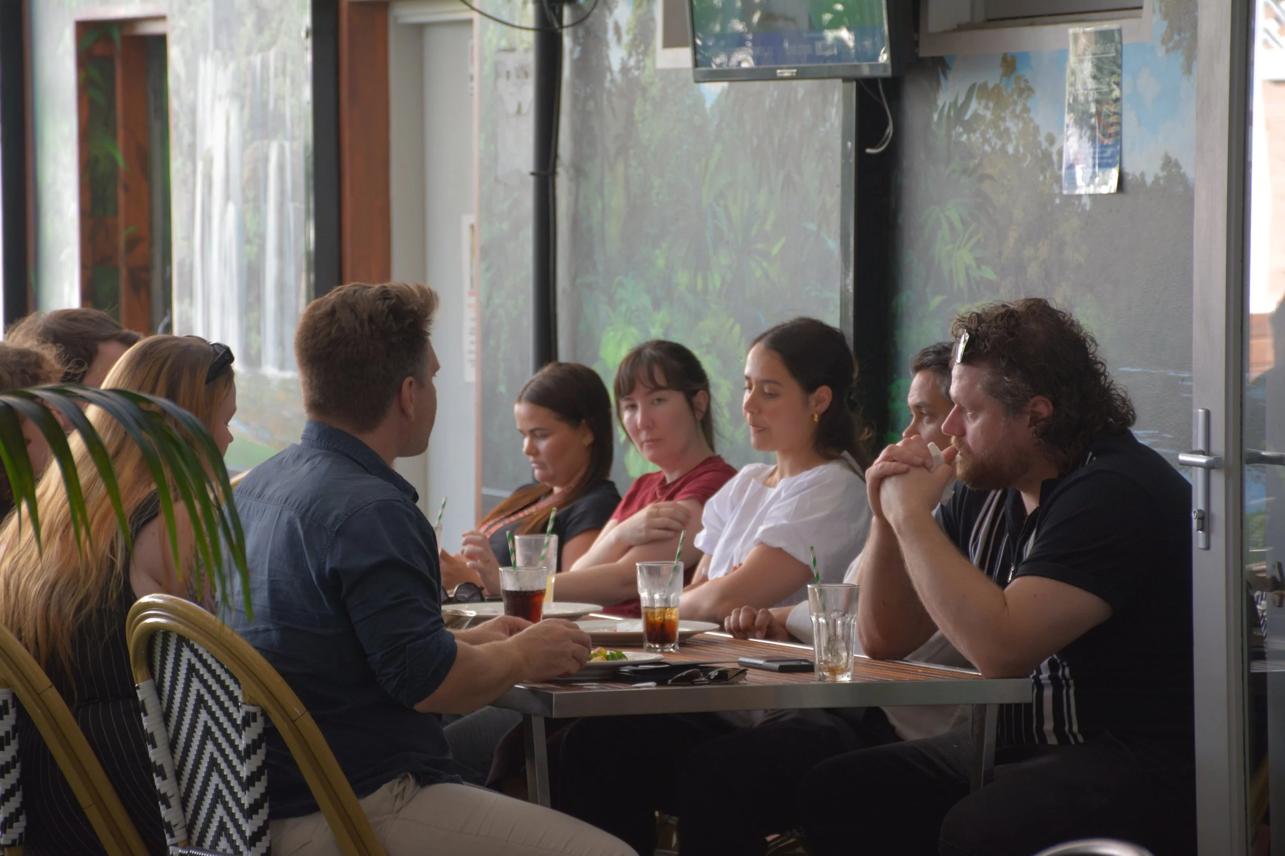 A group of people sitting at a long table in a room with large windows and a painted mural of nature on the wall, having drinks and talking.