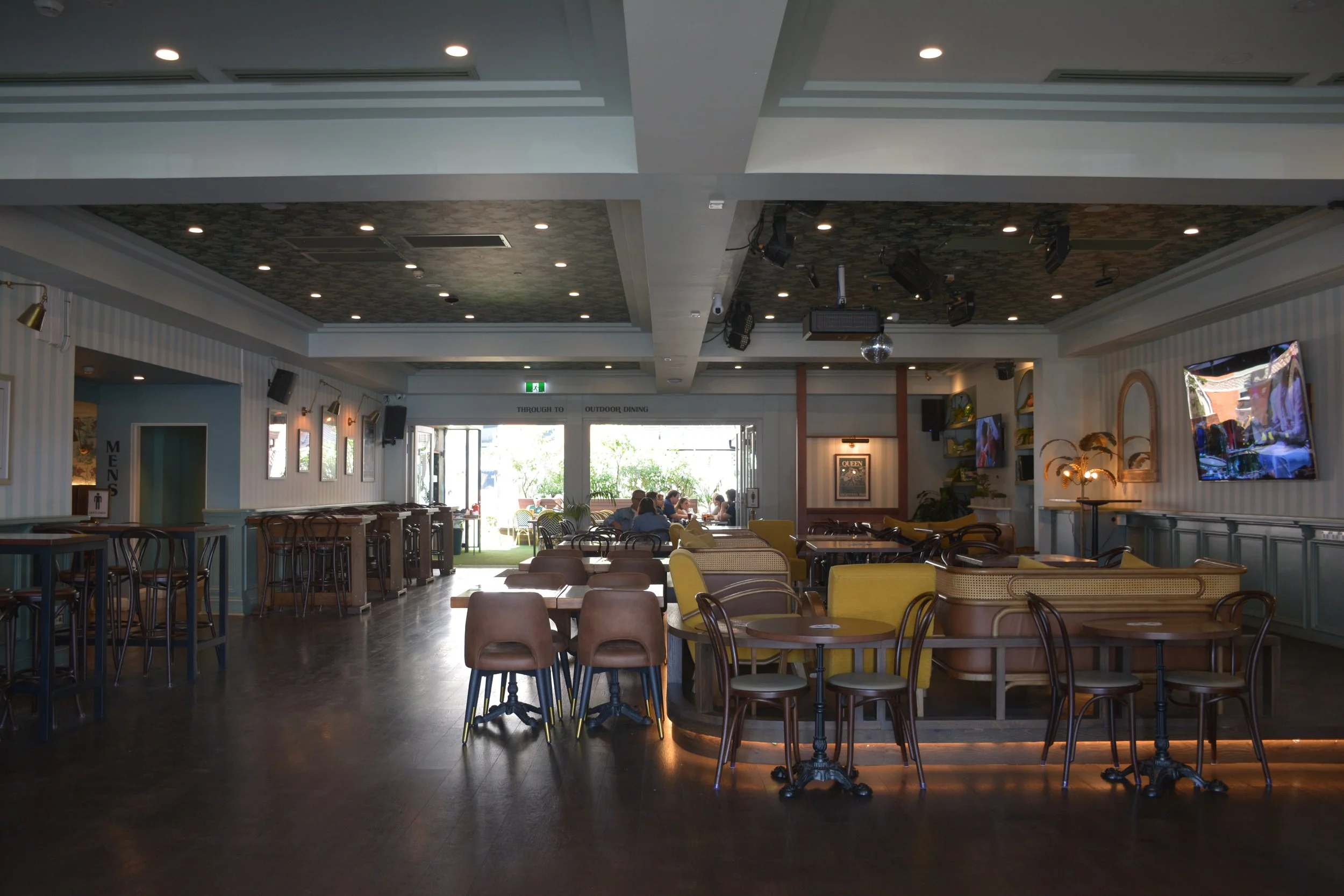 Interior of a restaurant or cafe with tables and chairs, some people dining, large windows letting in natural light, multiple televisions on the walls, and a ceiling with recessed lighting.