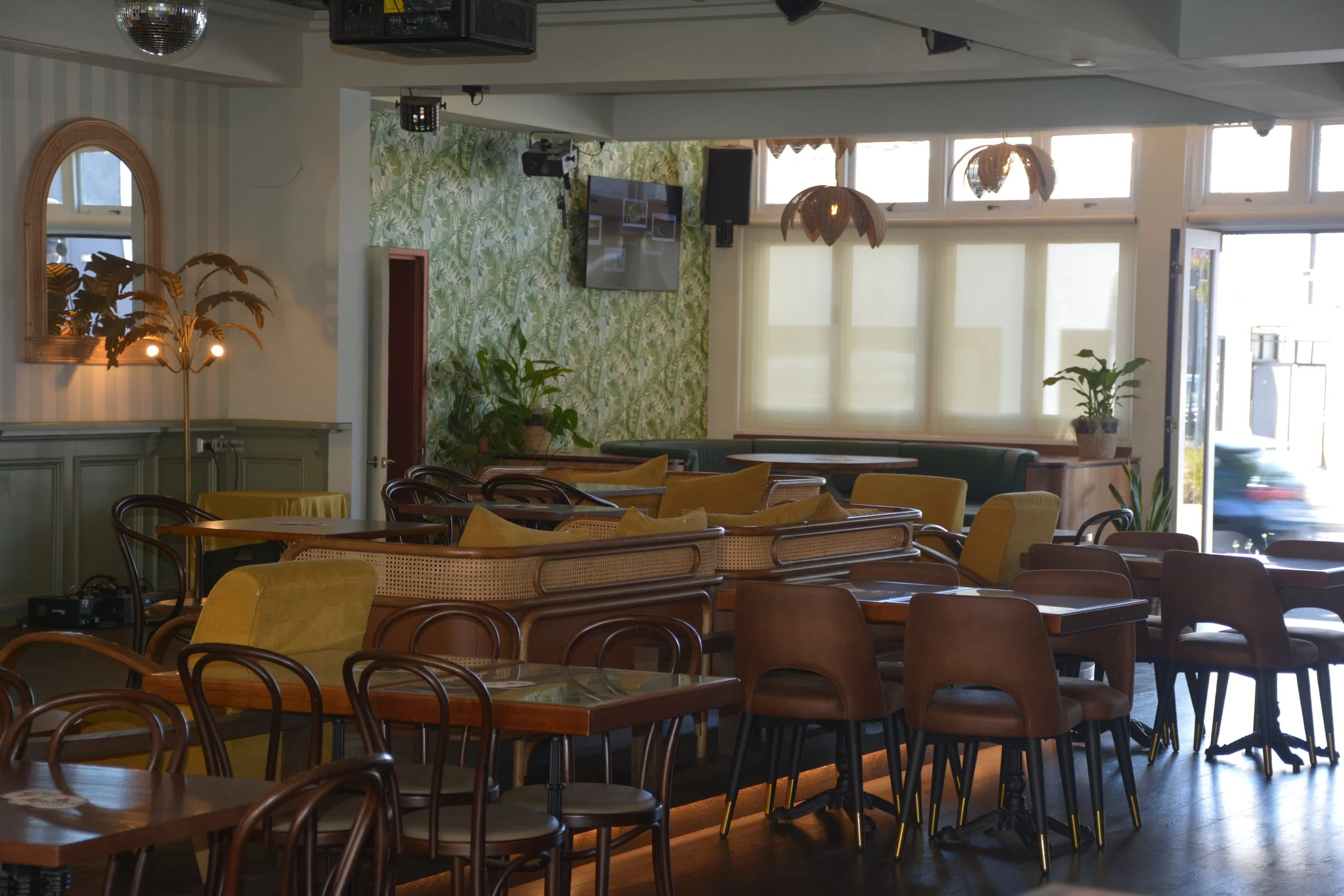 Interior of a cozy restaurant or cafe with wooden tables and chairs, decorated with green plants and botanical wallpaper, large windows letting in natural light, and tropical pendant lights hanging from the ceiling.