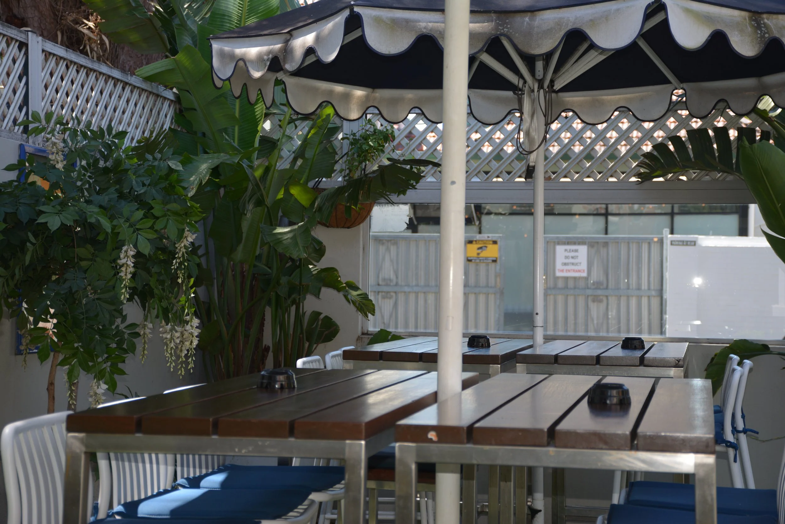 Outdoor dining area with wooden tables and white chairs, surrounded by green leafy plants, including large banana leaves, with a canopy providing shade.
