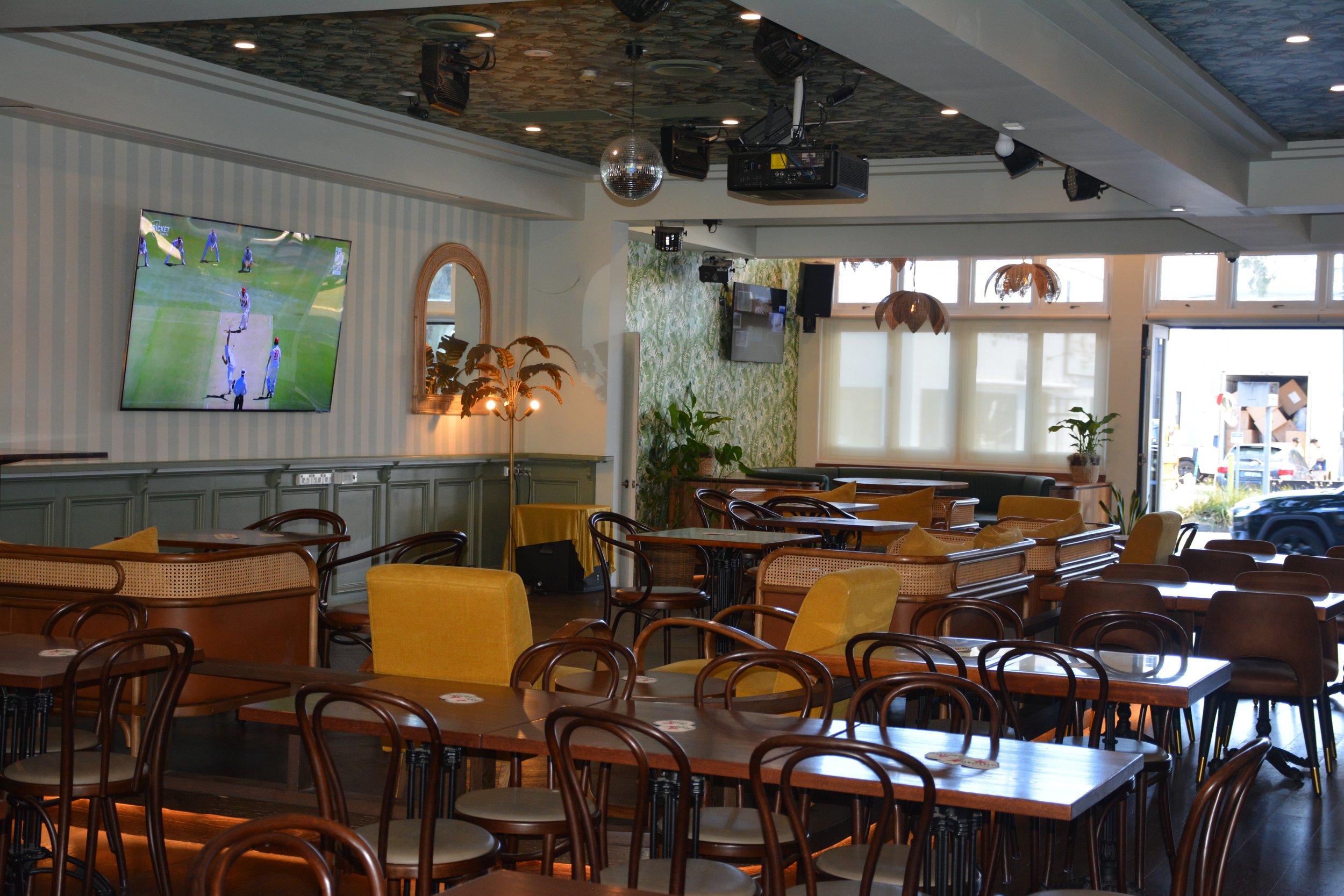 Interior of a restaurant or cafe with wooden tables and chairs, large windows letting in natural light, and a TV displaying a cricket match on the wall. Decor includes potted plants, decorative light fixtures, and a mirror on the wall.