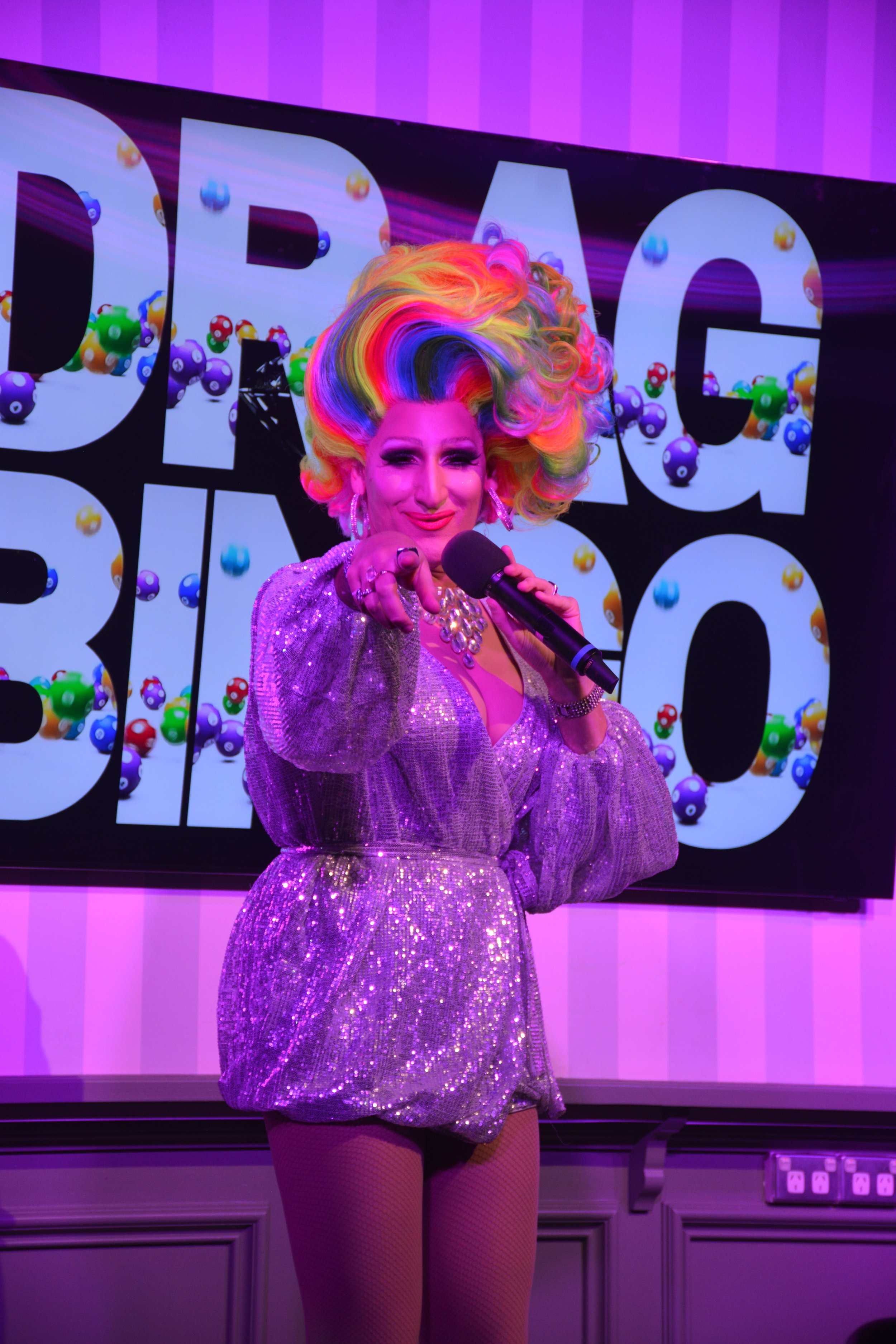 A drag queen with rainbow hair, wearing a sparkly purple outfit, pointing and smiling at the camera, standing in front of a background with large letters and colorful spheres.