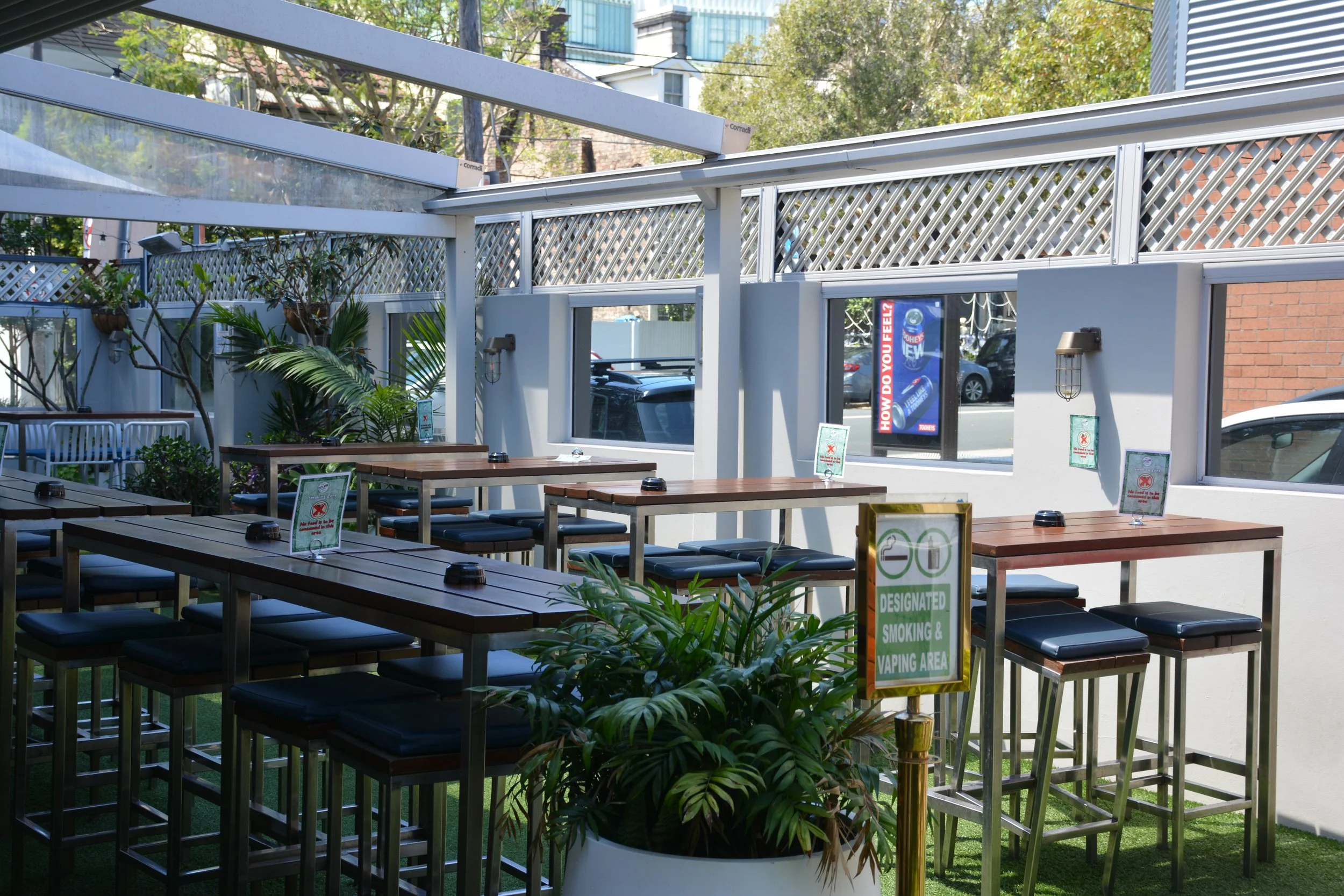 Outdoor patio seating with wooden tables and black cushioned chairs, surrounded by green plants and a white lattice fence, with signs indicating designated smoking and vaping area.