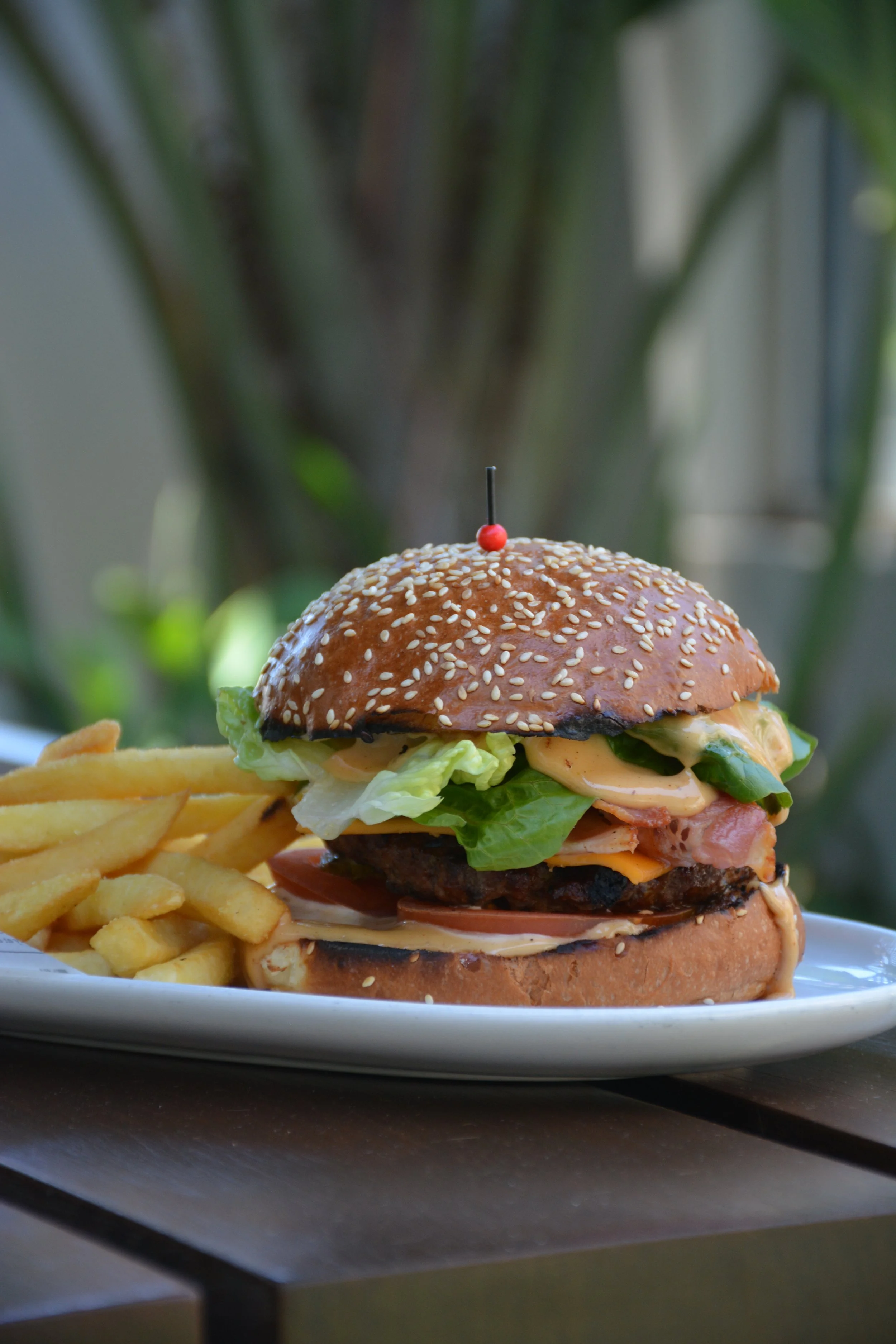 A cheeseburger with a sesame seed bun, lettuce, tomato, cheese, bacon, and a beef patty, served with French fries on a white plate at an outdoor setting.