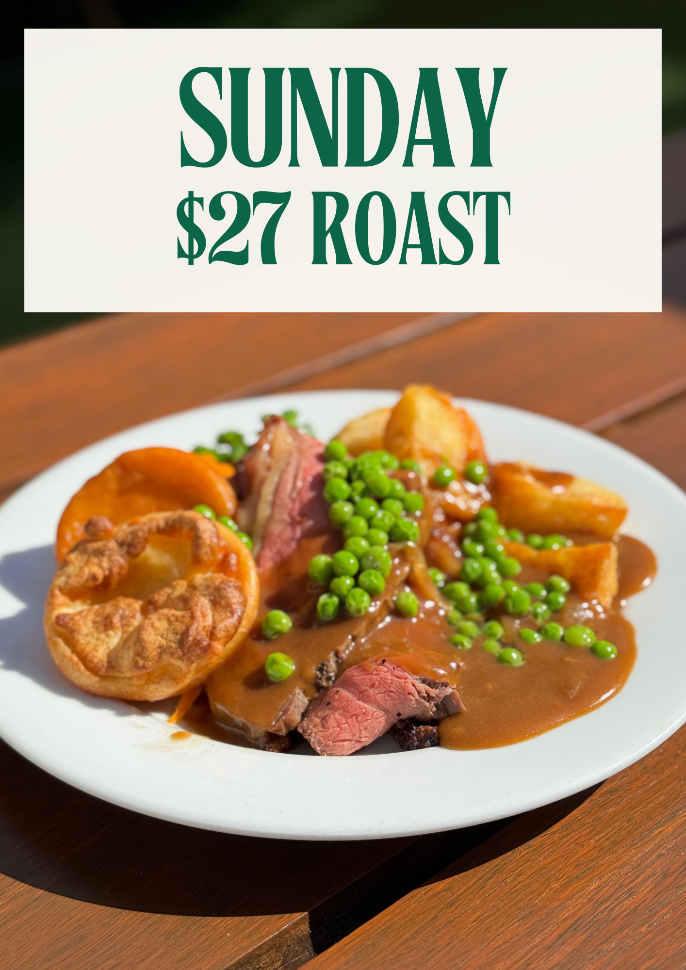 A white plate of roast beef with gravy, green peas, roasted potatoes, and a Yorkshire pudding on a wooden table.