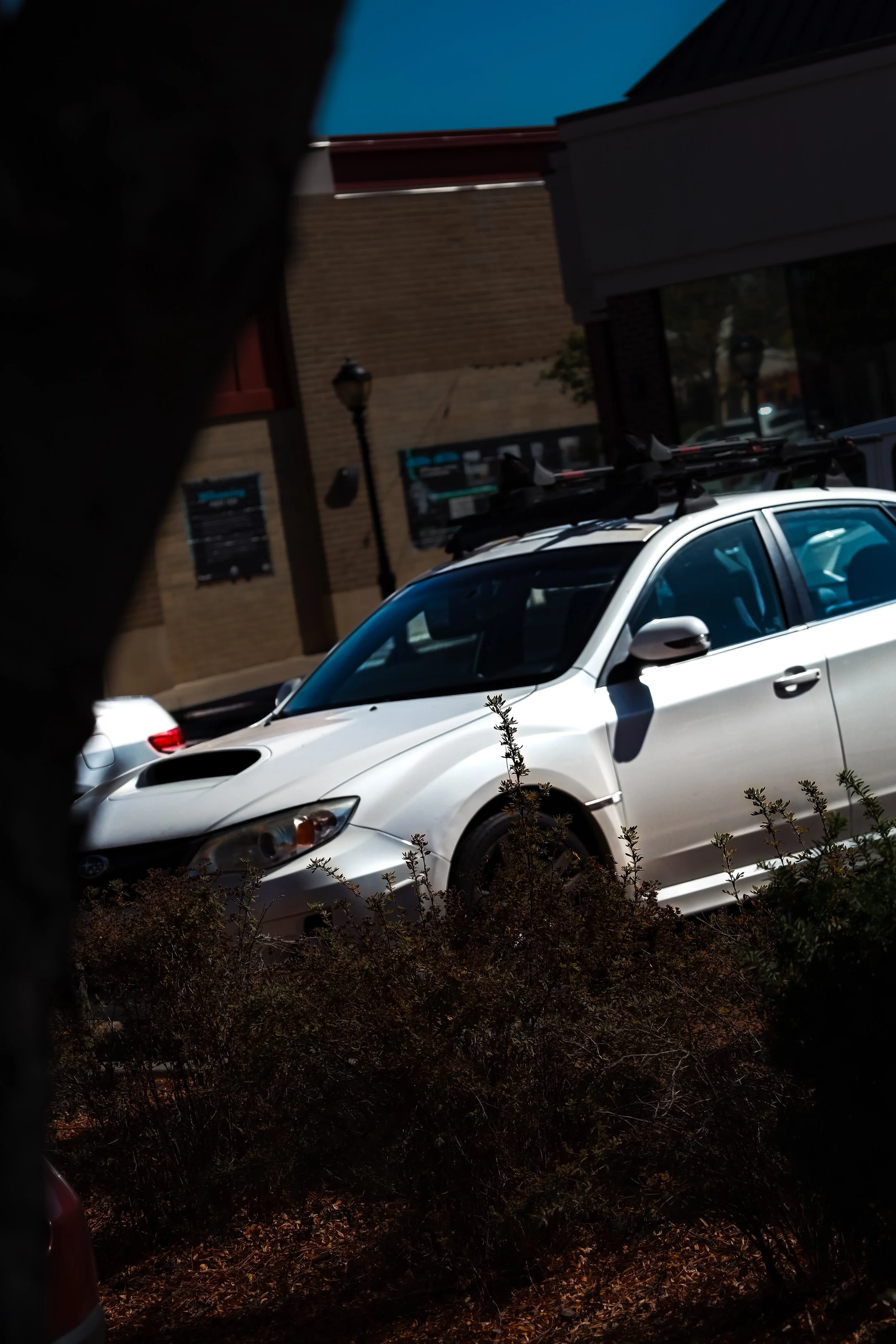 White coupe car parked outdoors near a sidewalk with bushes in the foreground, brick building, street lamp, and another vehicle in the background.