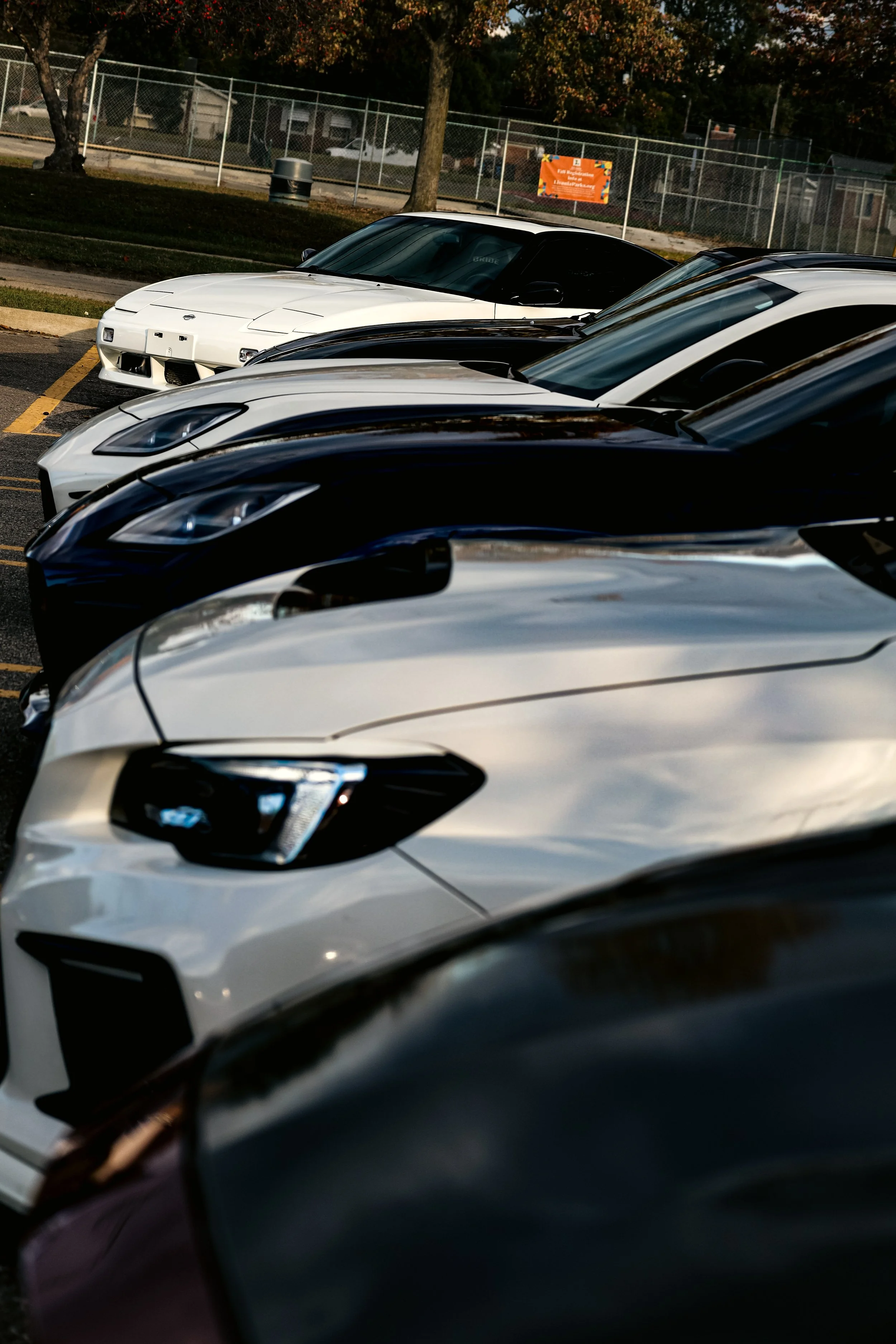 Several parked cars in a parking lot with a chain-link fence and trees in the background, during daylight.