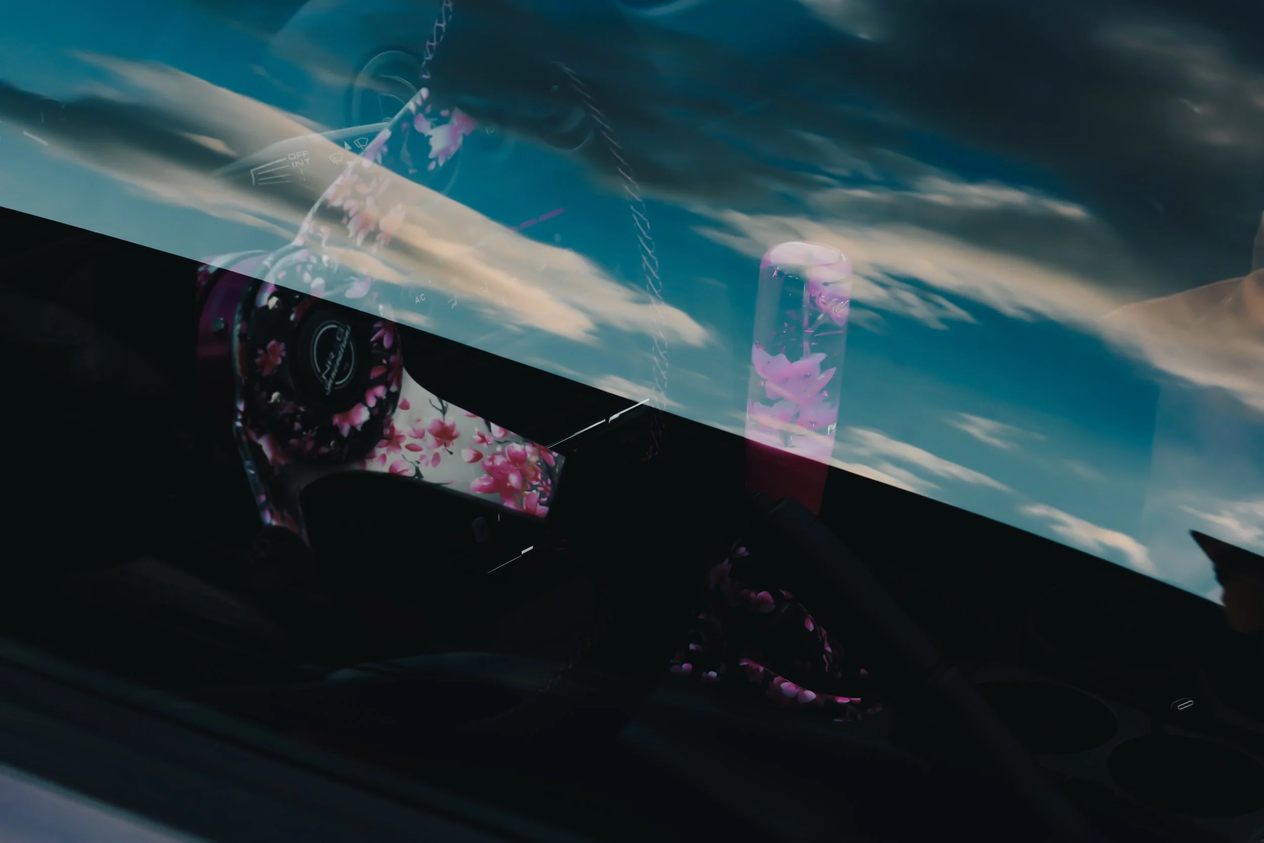 Interior view of a car with a black dashboard and a floral-patterned steering wheel cover, showing a reflection of a cloudy sky on the windshield.