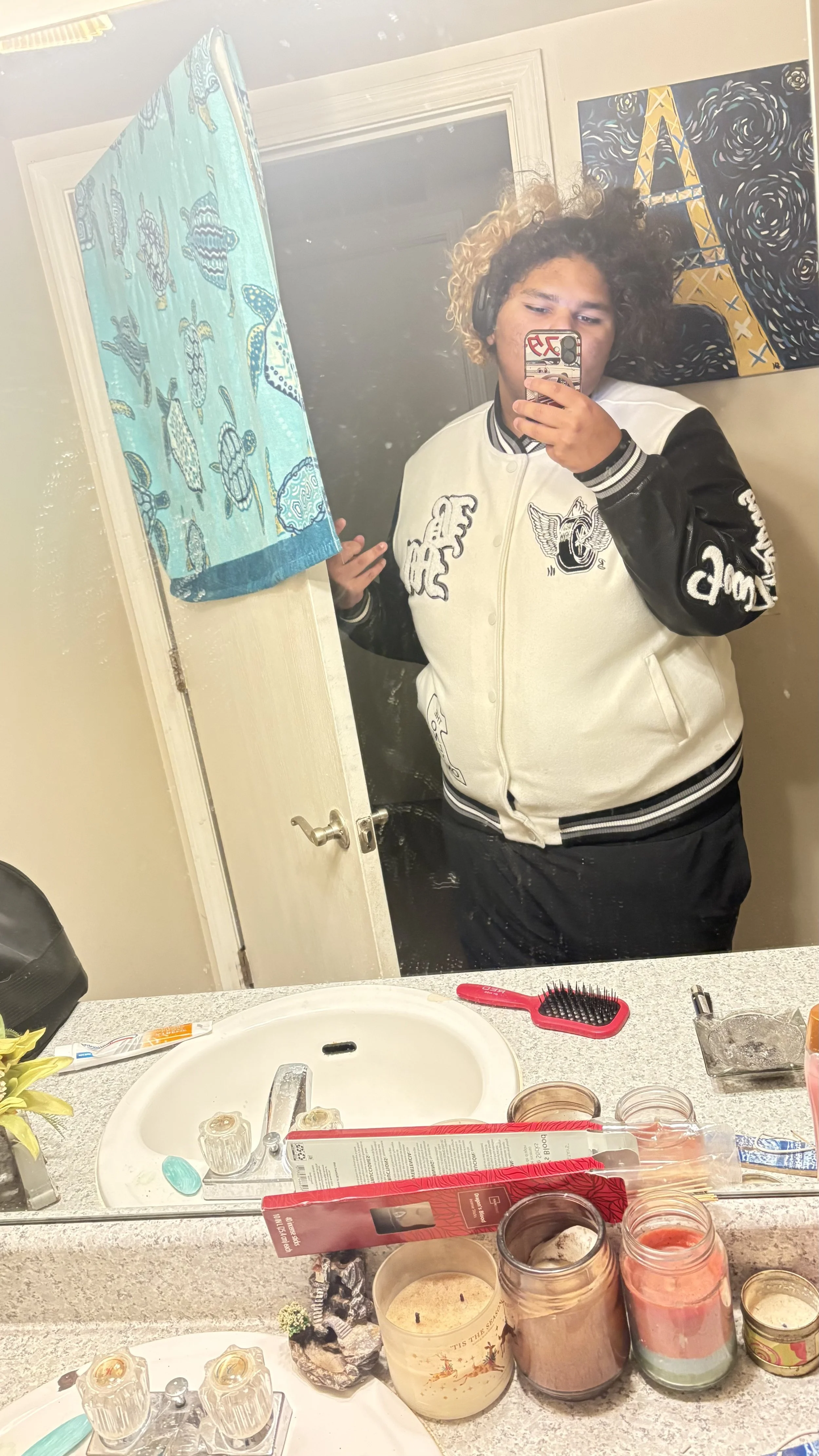 A person taking a mirror selfie in a bathroom, wearing headphones and a varsity jacket, with a colorful wall and painting of the Eiffel Tower in the background. The bathroom counter has various candles, toiletries, and a red hairbrush.