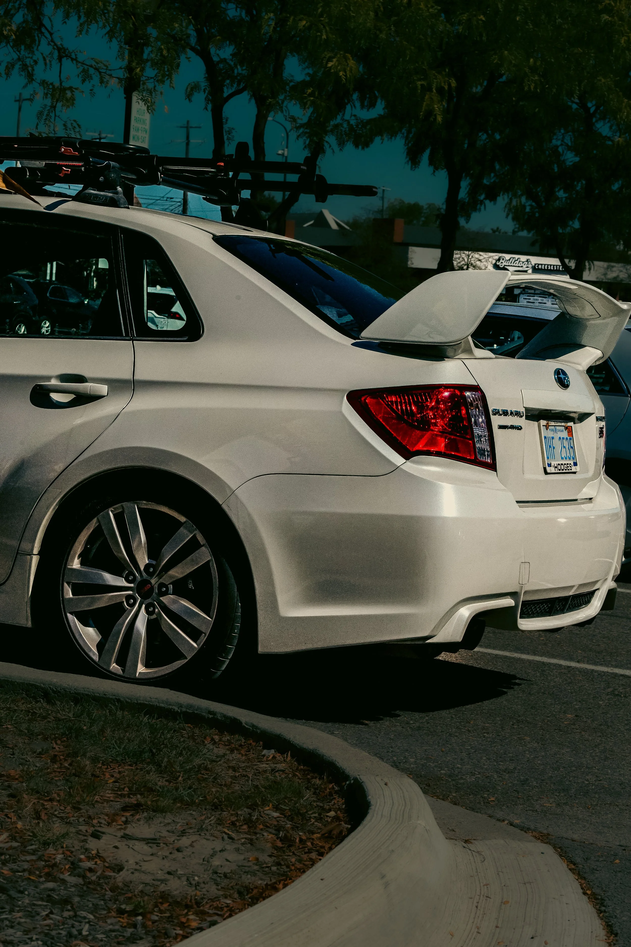 A white Subaru car parked in a parking lot with a roof rack and a large rear spoiler. The car's license plate is visible. There are trees and a building in the background. The parking lot has a curved concrete curb and some fallen leaves.