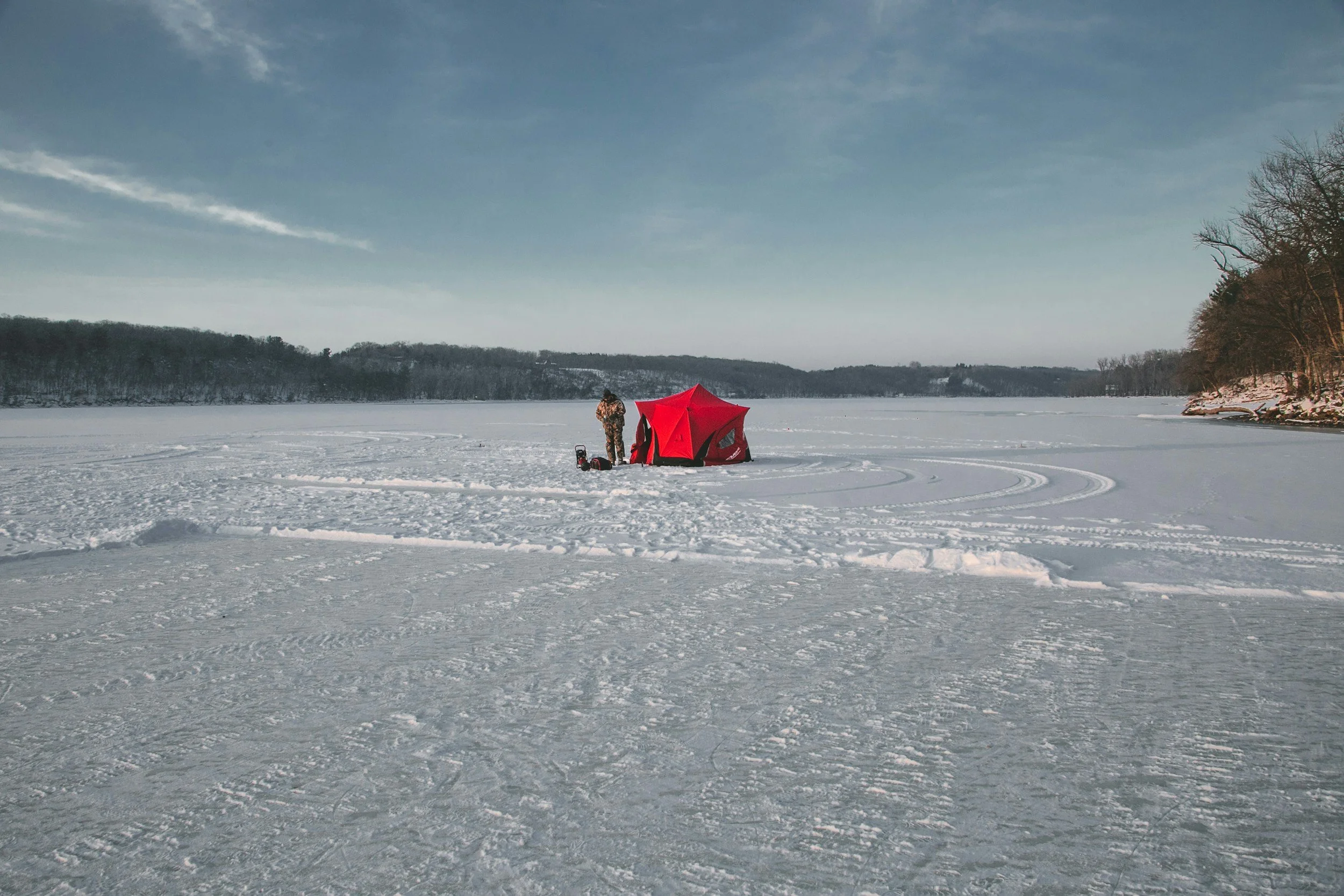 A person dressed in camouflage clothing standing next to a red tent on a snow-covered lake, with snow-covered hills and trees in the distance under a cloudy sky.
