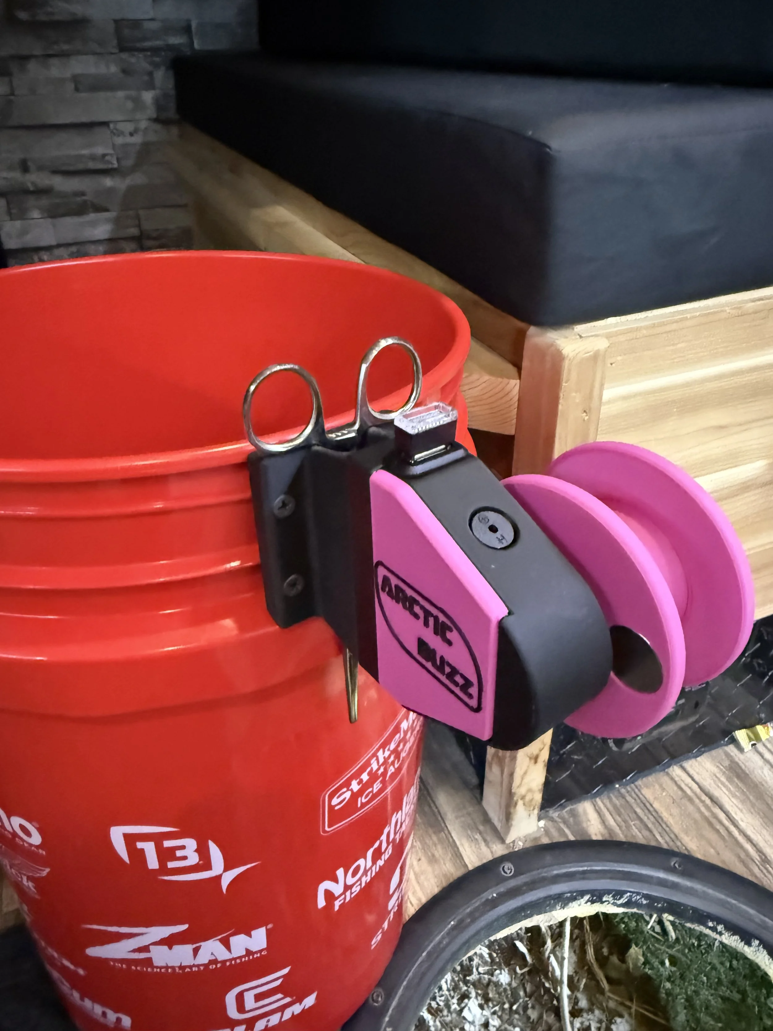 A red plastic bucket with white graphics and a black handle, a pink plastic bucket, and a black tool with a metal ring attached, all placed on a service cart with wooden tiers and metal edges, next to a bicycle wheel with a black rubber tire and spok