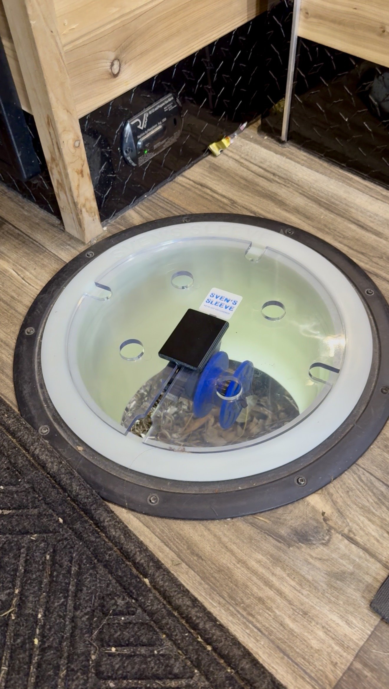 A ceiling host with a recessed lighting fixture in a room with wood flooring and shelving. The fixture has a black cover and is secured by screws.