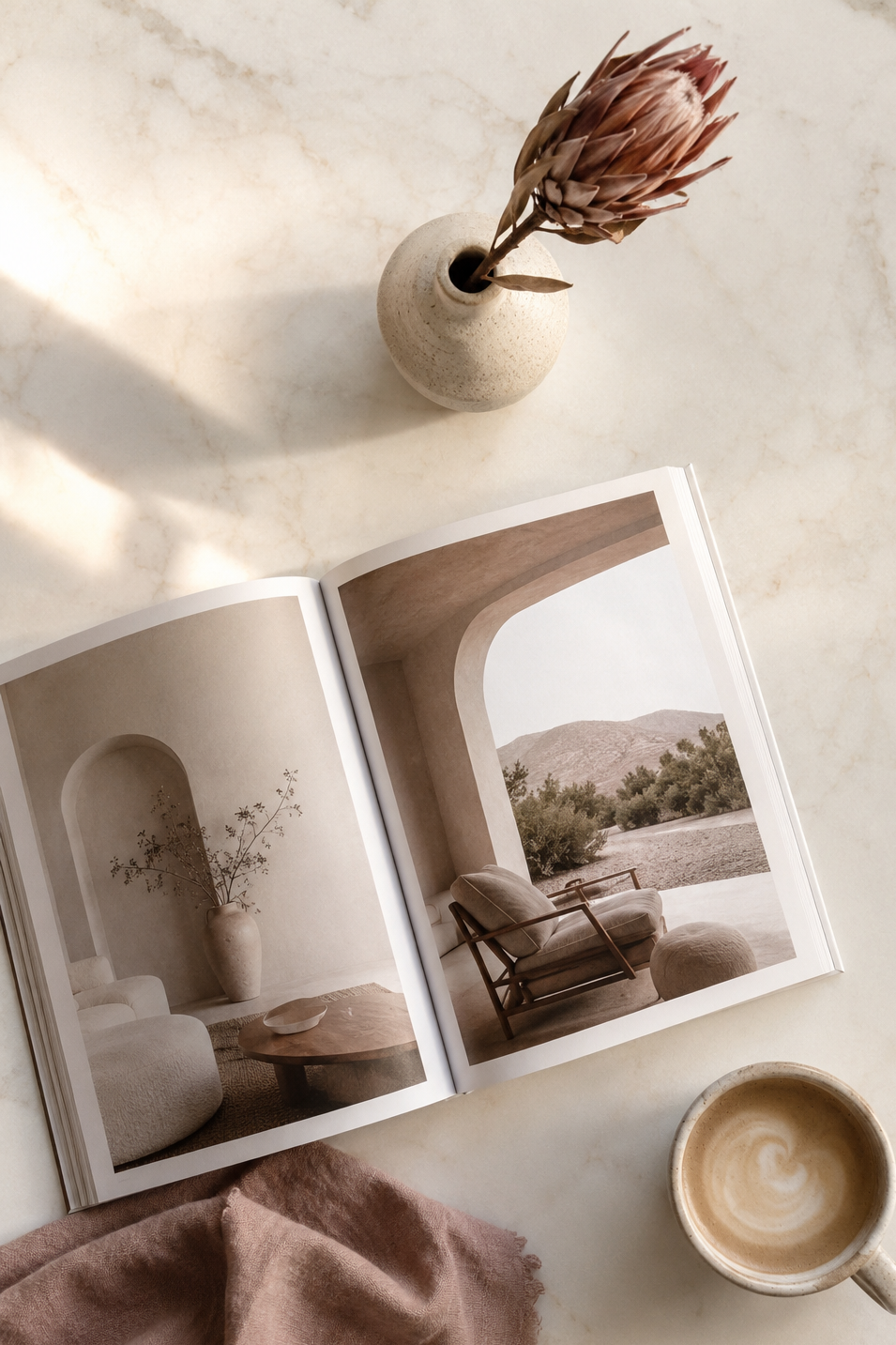 Open magazine on a marble surface displaying images of modern interior architecture, with a small ceramic vase holding a dried flower, a cup of coffee with foam, and a pink cloth nearby.