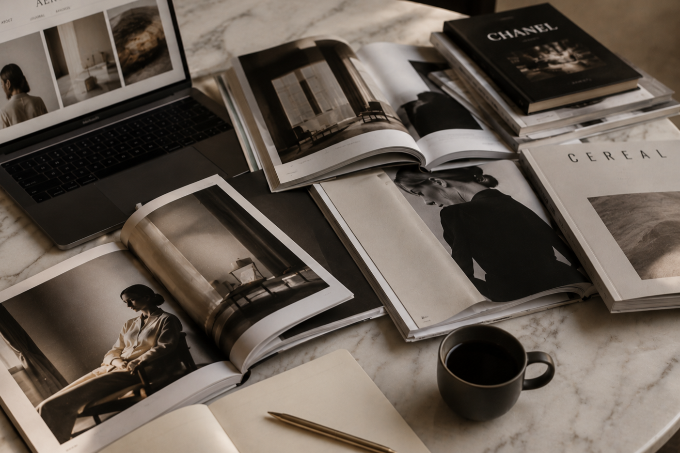 Multiple open magazines with black and white photographs of a woman, a laptop displaying images, a closed book titled 'CHANEL,' another magazine titled 'CEREAL,' a cup of coffee, and a blank notebook with a pen on a marble table.