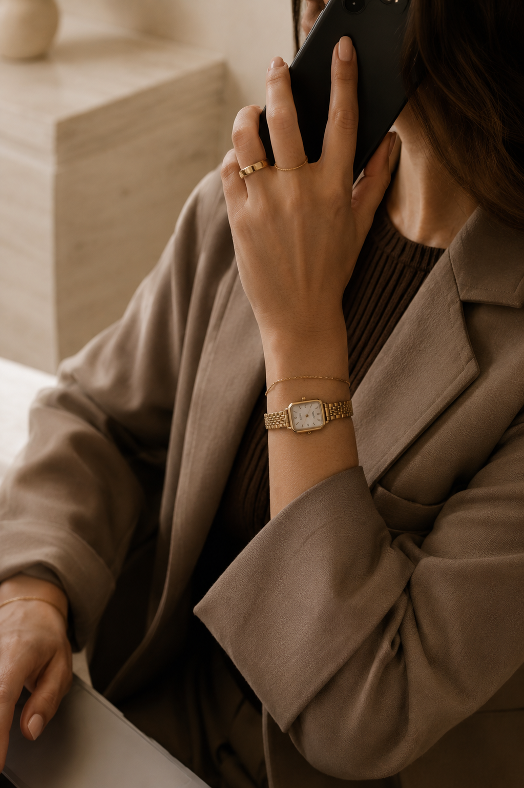 A woman in a beige blazer and brown top is talking on the phone while sitting at a table, wearing a gold wristwatch, rings, and a delicate bracelet.