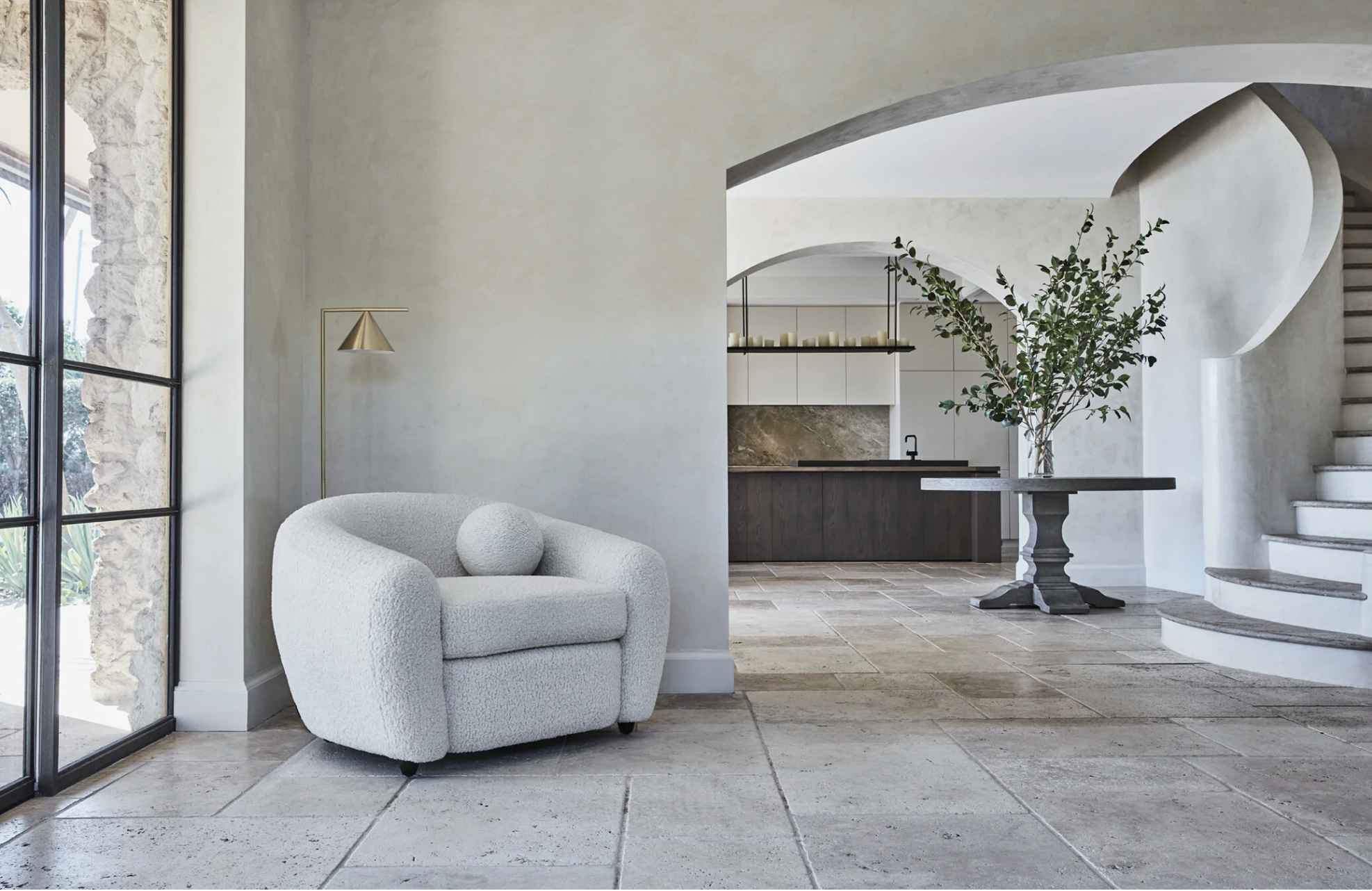 Coco Republic catalogue featuring the interior of a modern house with a white armchair near large windows, a dark wood table with a tall vase and greenery, stone tile flooring, and a staircase with curved walls and railing.
