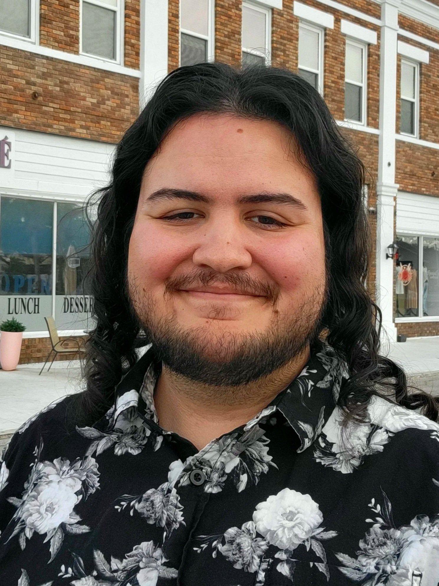 A person with long black hair, a beard, and a mustache taking a selfie outdoors in front of a brick building with white-framed windows. The person is smiling and wearing a black shirt with a white floral pattern.