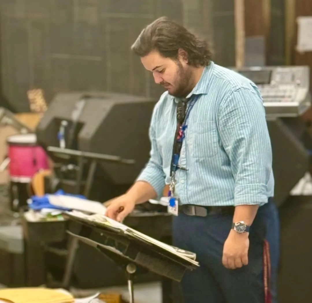 A man with dark, wavy hair and a beard, wearing a light blue button-up shirt and a watch, stands at a music stand with a book or sheet music on it, in a cluttered room with musical equipment and supplies in the background.