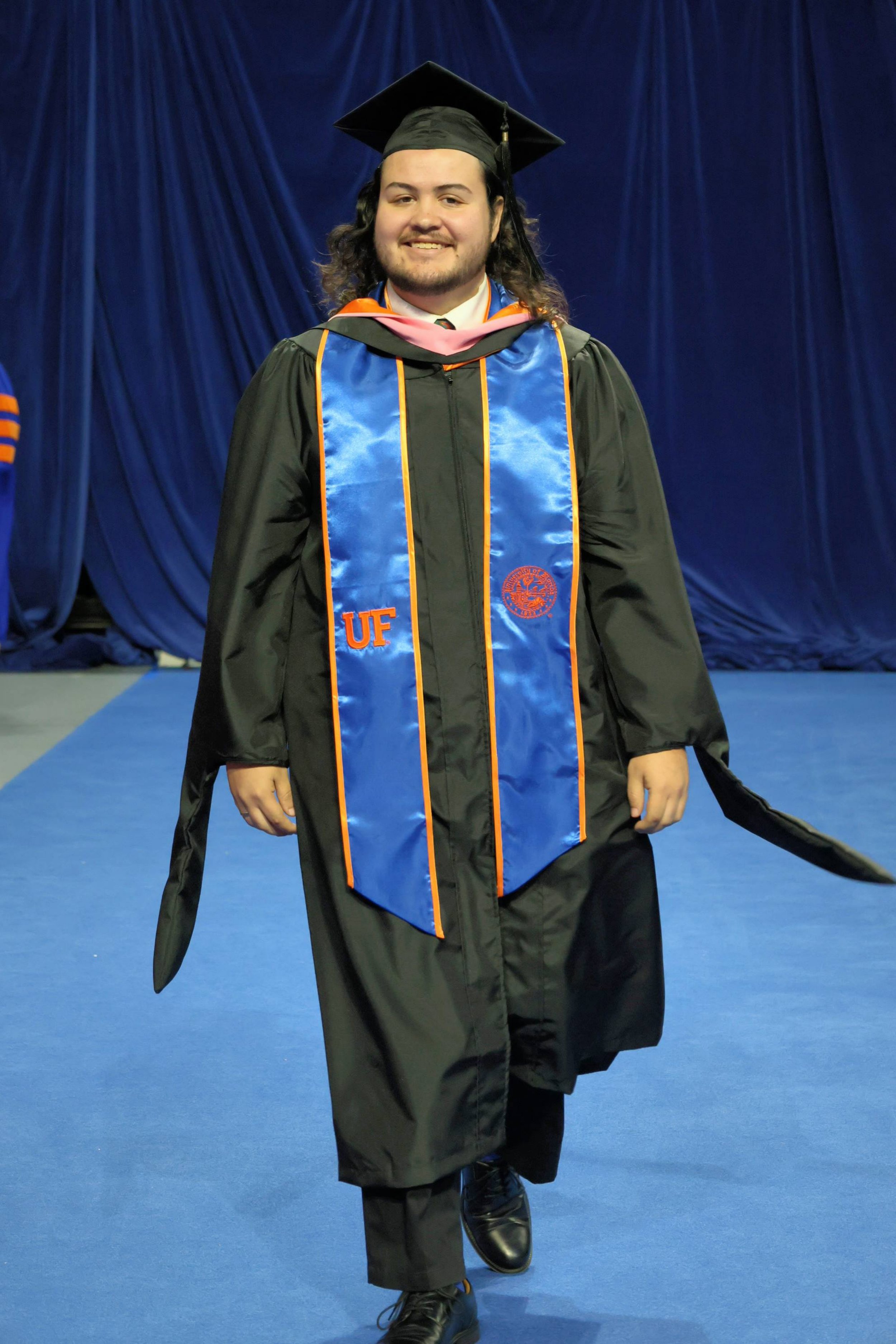 A young man in a black graduation gown and cap, with a blue and orange stole, is smiling and walking on a blue carpet in front of a dark blue curtain.