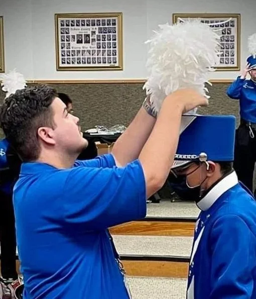 A young man in a blue shirt helps a younger person in a blue dress uniform and tall blue shako hat adjust a feathered plume during a ceremony or parade in a room with framed photos on the wall.