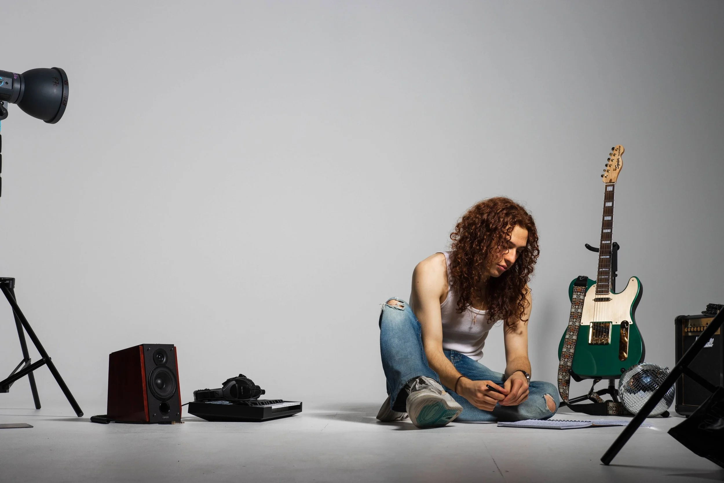 Michael Ebejer, a man with curly red hair sitting on the floor in a music studio, surrounded by musical equipment, including an electric guitar, speakers, and a keyboard. He is wearing a white tank top and ripped jeans, looking down at a book.