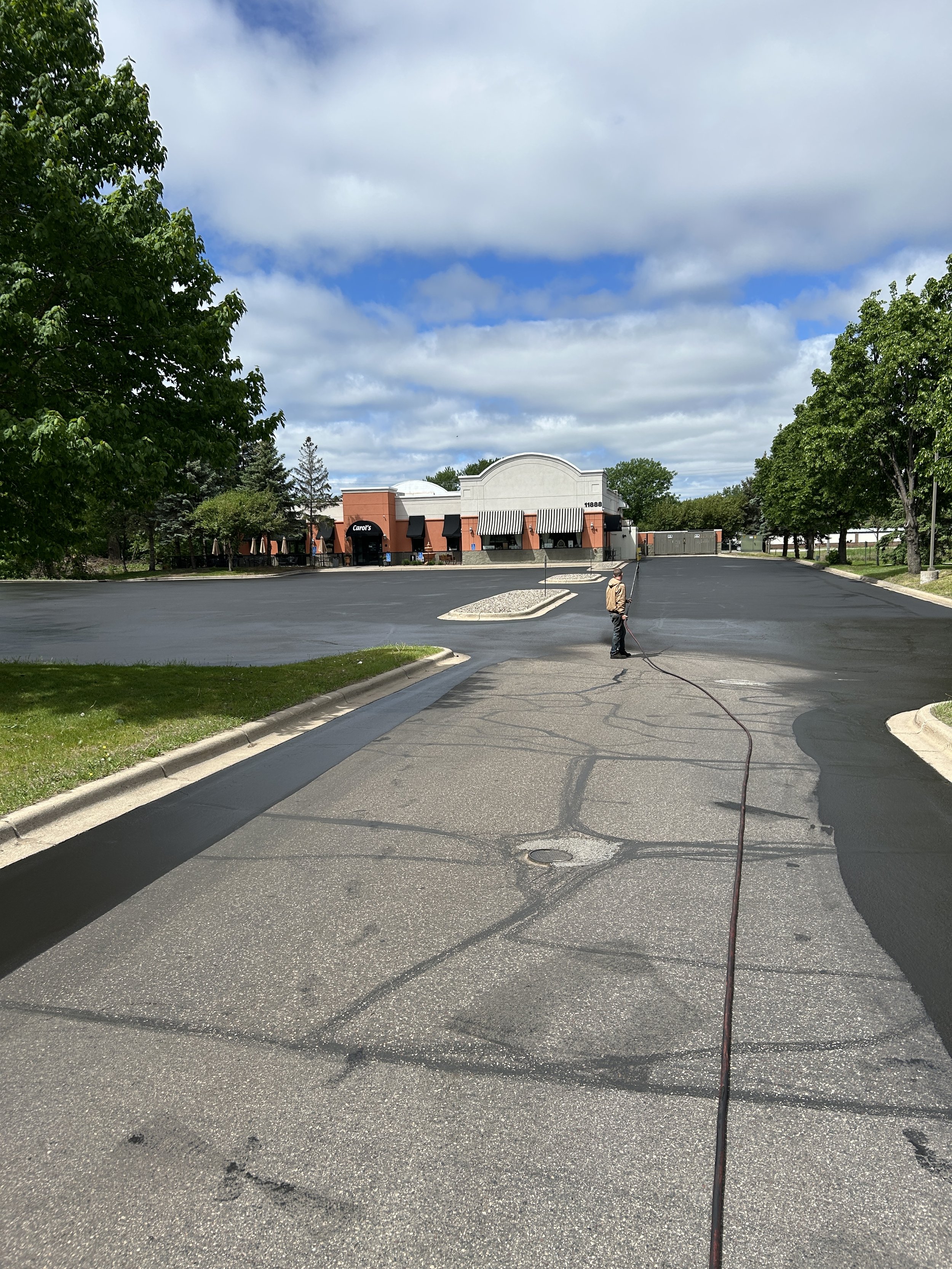 A 7 County Sealcoating professional applying a fresh asphalt sealcoat to a commercial parking lot after completing crack filling and asphalt repair at Carol's Restaurant in Blaine, Minnesota.