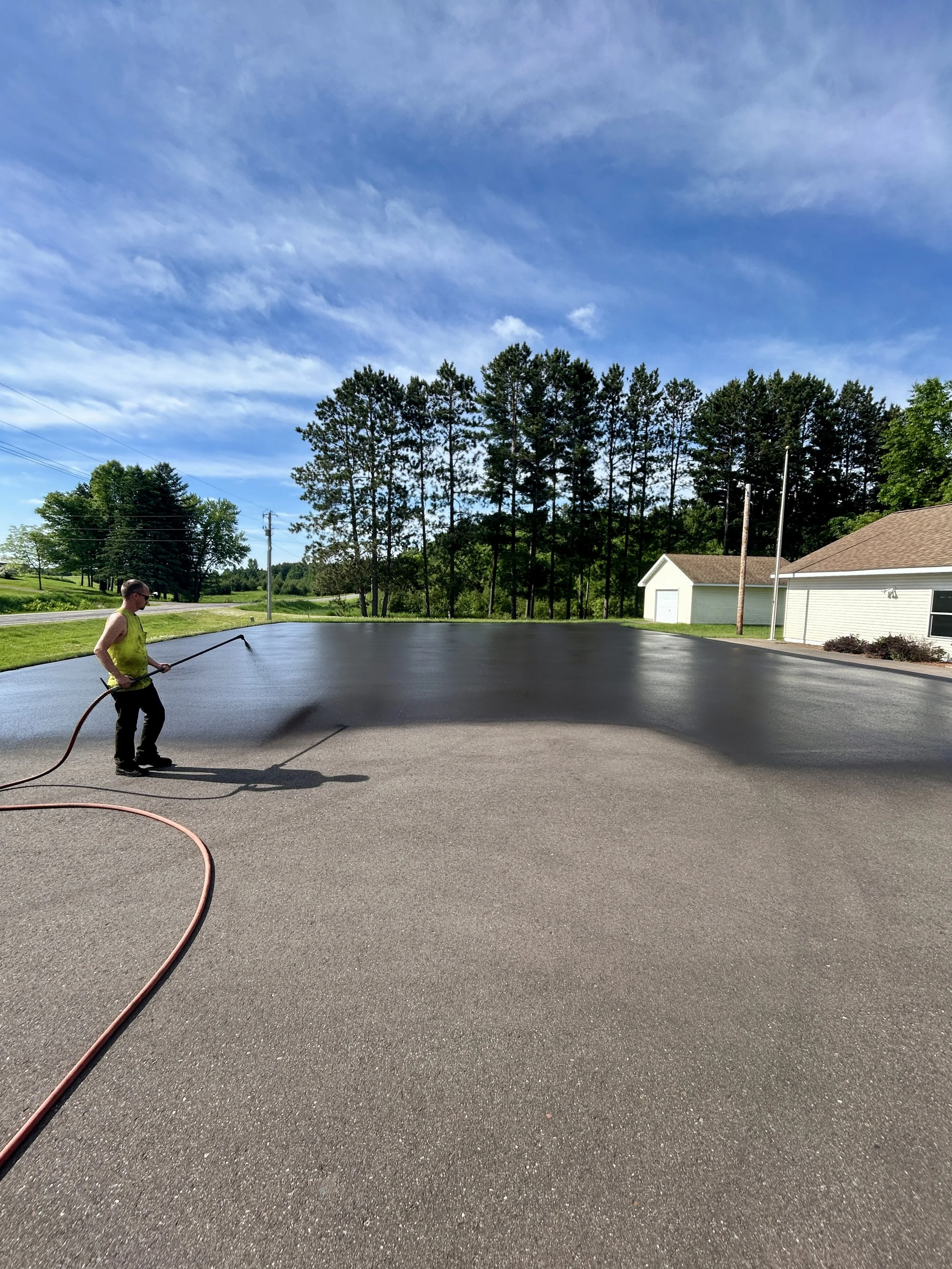 A 7 County Sealcoating professional applying a fresh asphalt sealcoat to a city municipal building parking lot in Isanti, Minnesota, after completing expert site prepping, cleaning, edging, and crack filling
