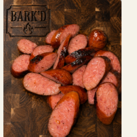 Sliced cooked sausage on a wooden cutting board with the word 'BARK'D' engraved in the top left corner.