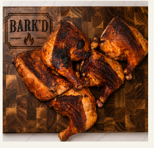 Four grilled chicken quarters on a wooden cutting board with the words 'BARK'D' and a flame logo engraved on it.