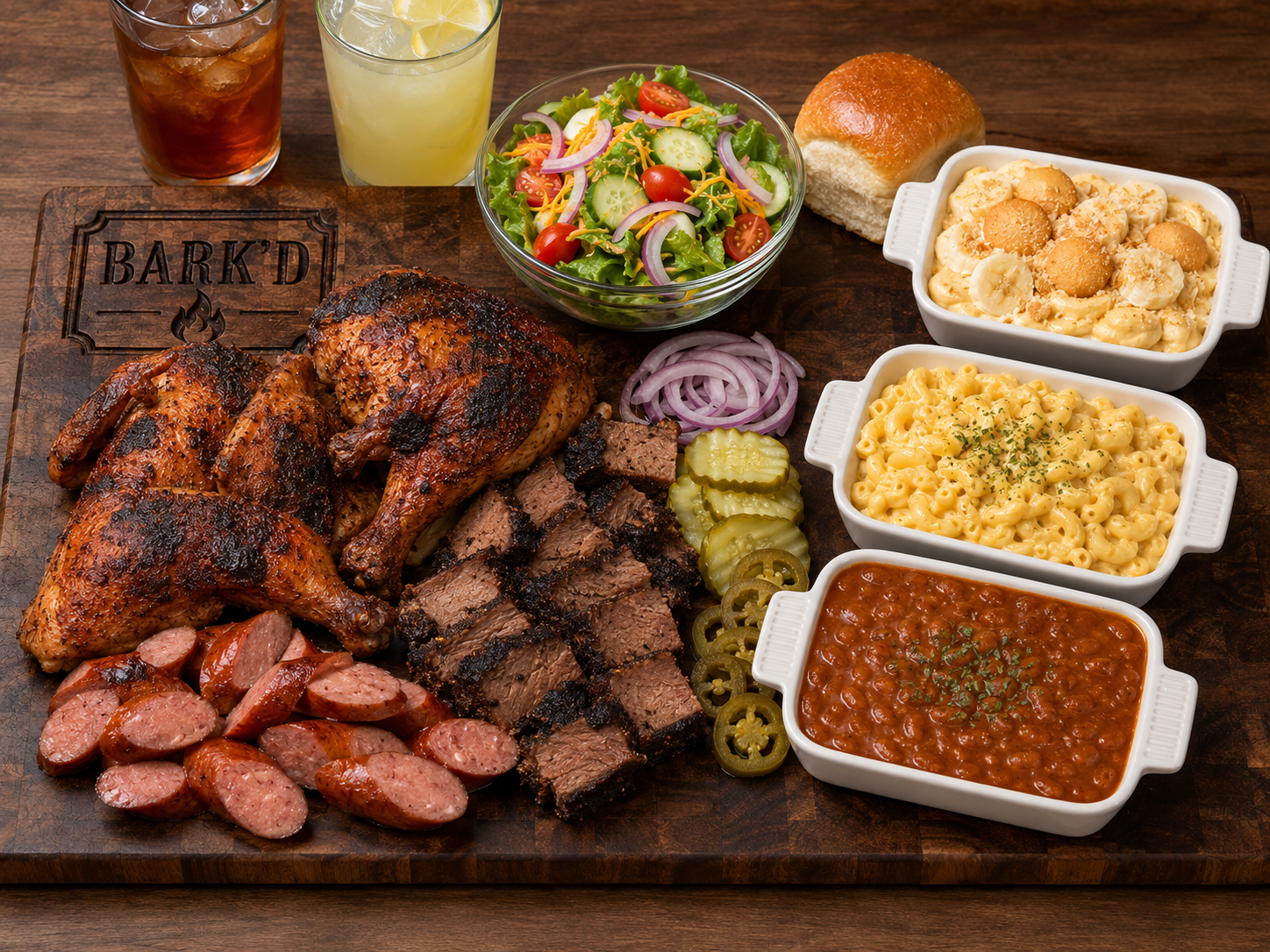 A platter of grilled meats including smoked sausage, grilled chicken, and brisket, served with sliced pickles and onions, alongside a salad, dinner roll, and three bowls of baked pasta and chili, with two glasses of iced tea and lemonade.