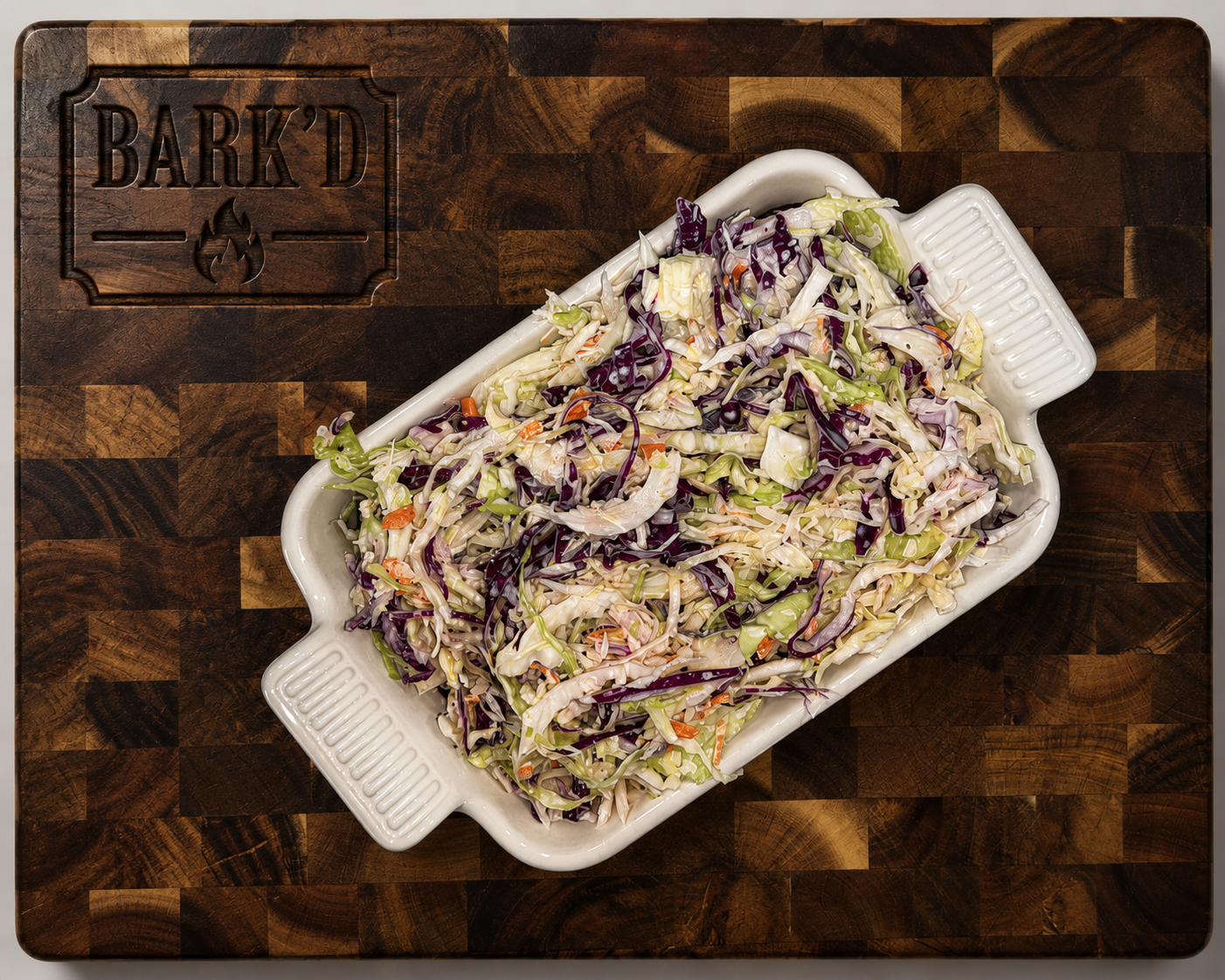 A rectangular white ceramic dish shaped like a pig, filled with coleslaw salad, placed on a wooden cutting board with a "BARK'D" logo and flame design.