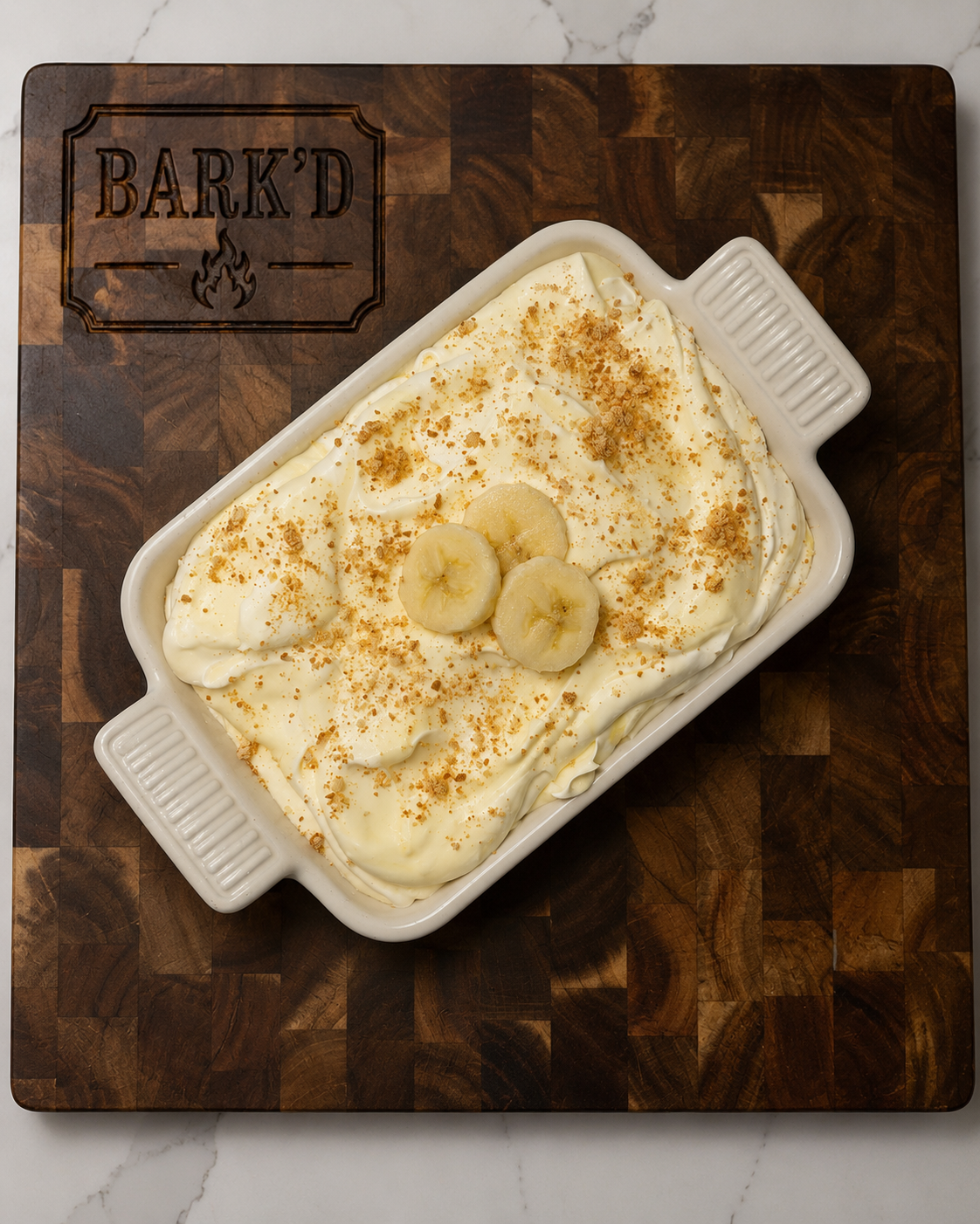 Banana pudding dessert topped with sliced bananas and crushed cookies in a white baking dish on a wooden cutting board.