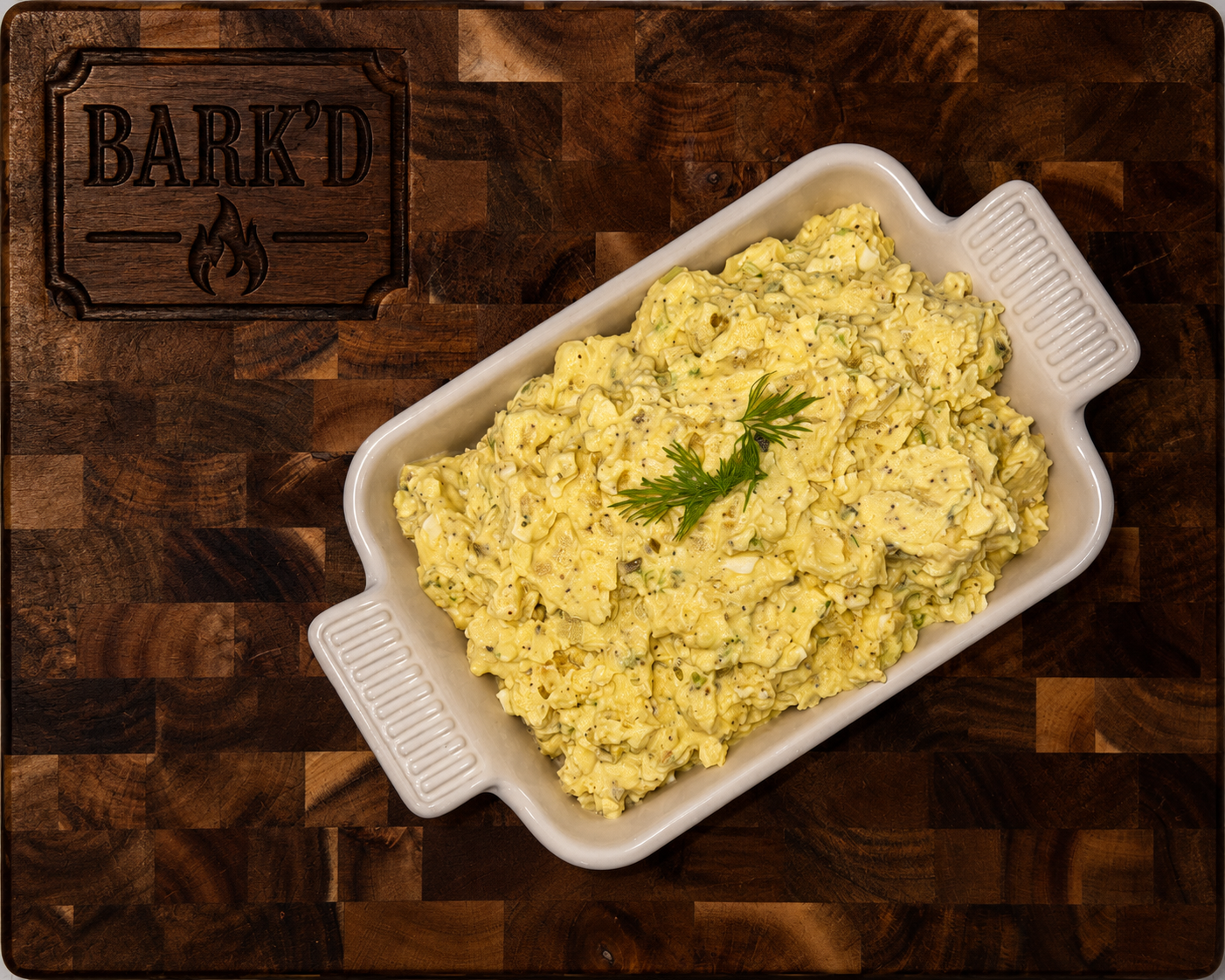 A white rectangular dish filled with creamy egg salad garnished with a sprig of fresh dill, placed on a dark wooden cutting board with a carved label that reads 'BARK'D'.