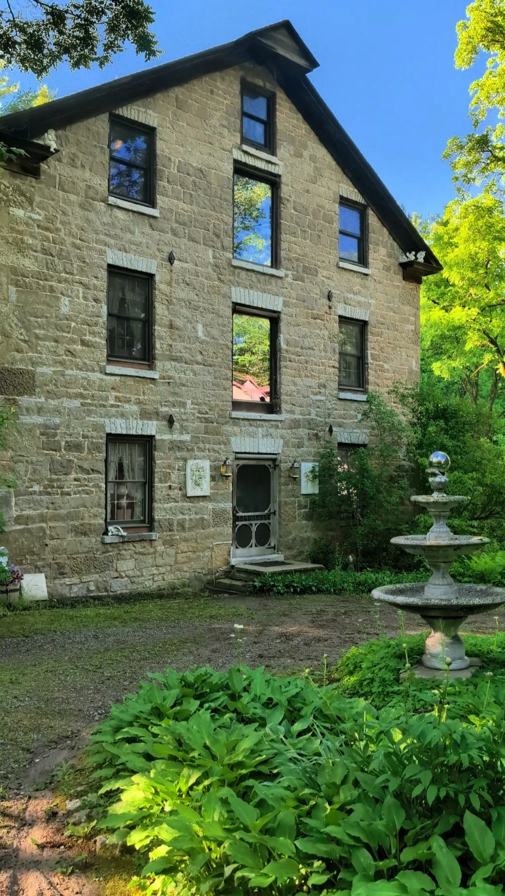 A stone house with four levels and black-framed windows, surrounded by green trees and bushes. There is a white decorative fountain in the front yard and a small garden to the left of the house.