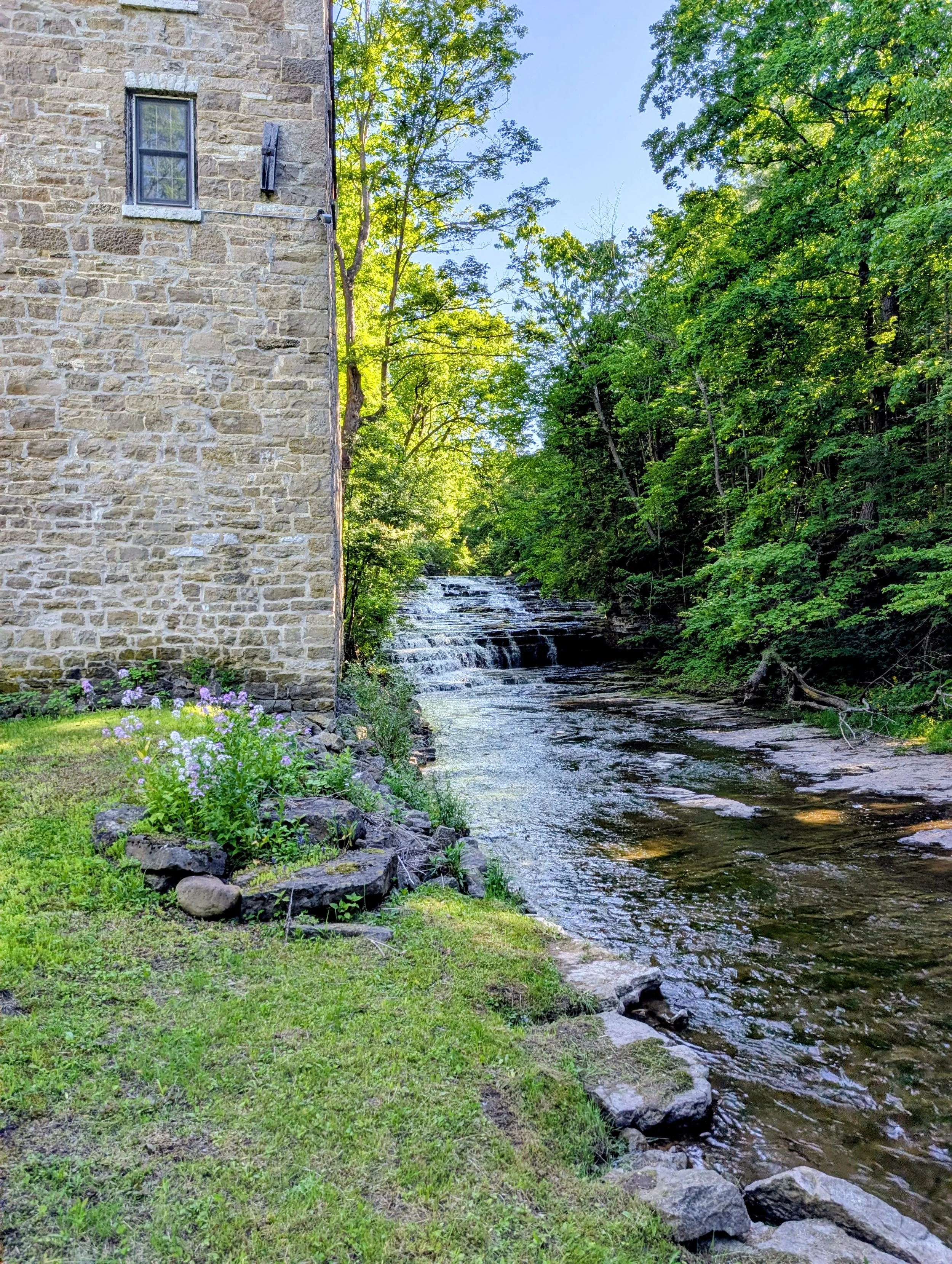 A stone building beside a creek surrounded by lush green trees and plants, with a small waterfall or cascade in the water.