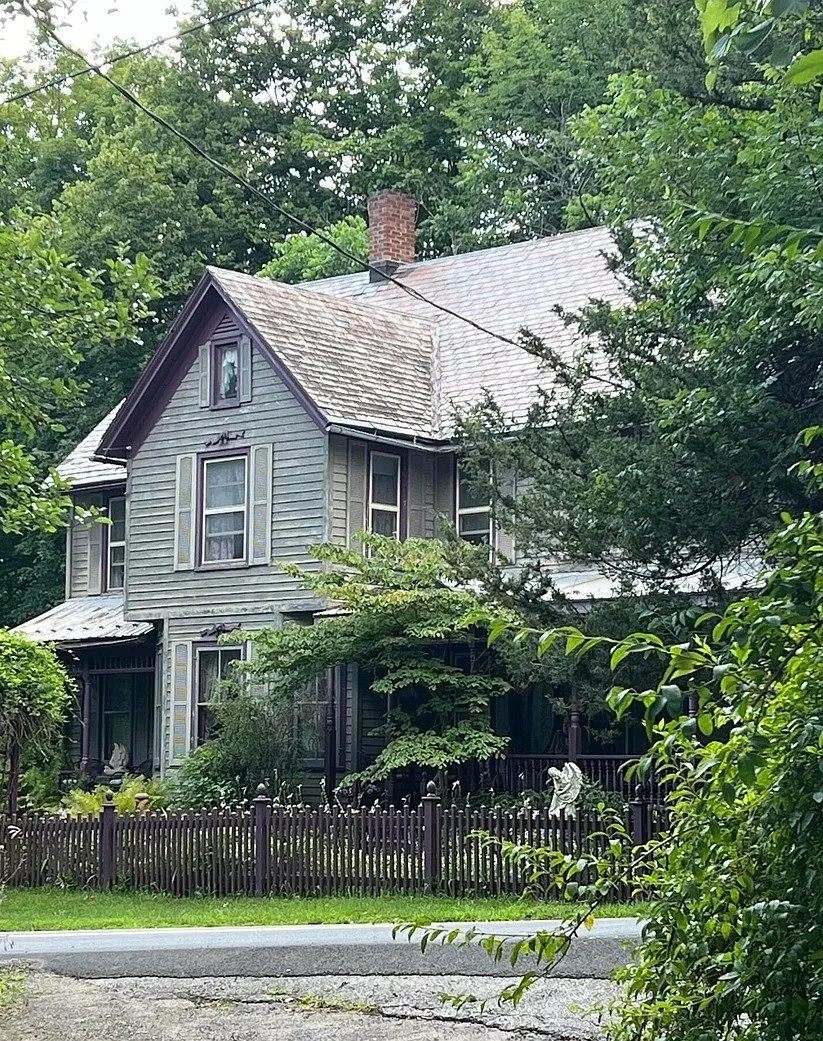 An old two-story house with weathered gray wooden siding, shrubs, a small tree in the front yard, a wooden picket fence, and surrounded by tall green trees, with a brick chimney and a sloped roof.