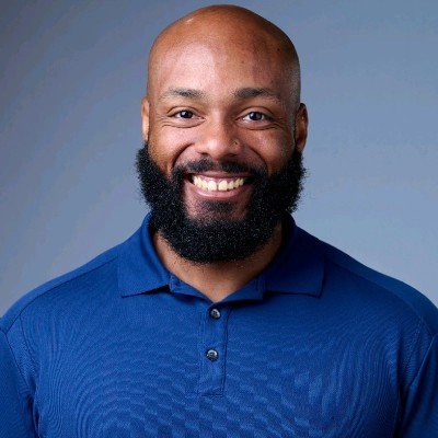 A smiling man with a beard and shaved head wearing a blue polo shirt against a gray background.