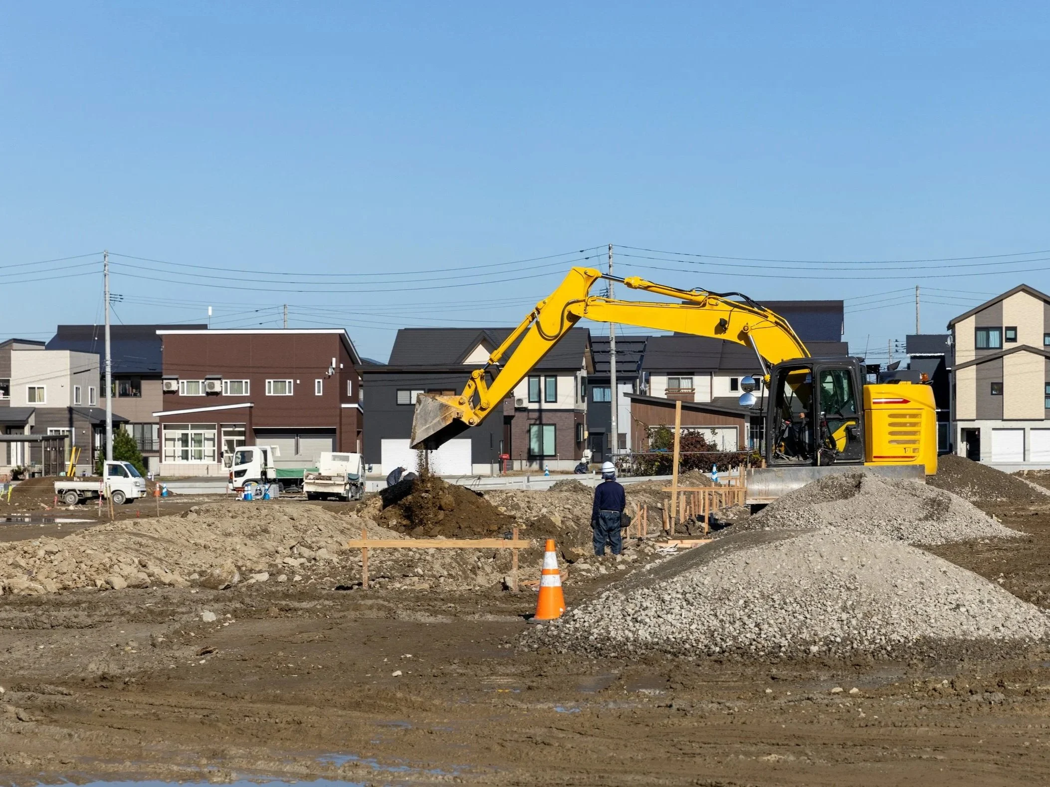 住宅地の建設工事で黄色いショベルカーが土を掘っている。周囲には家々が立ち並び、青空が広がっている。