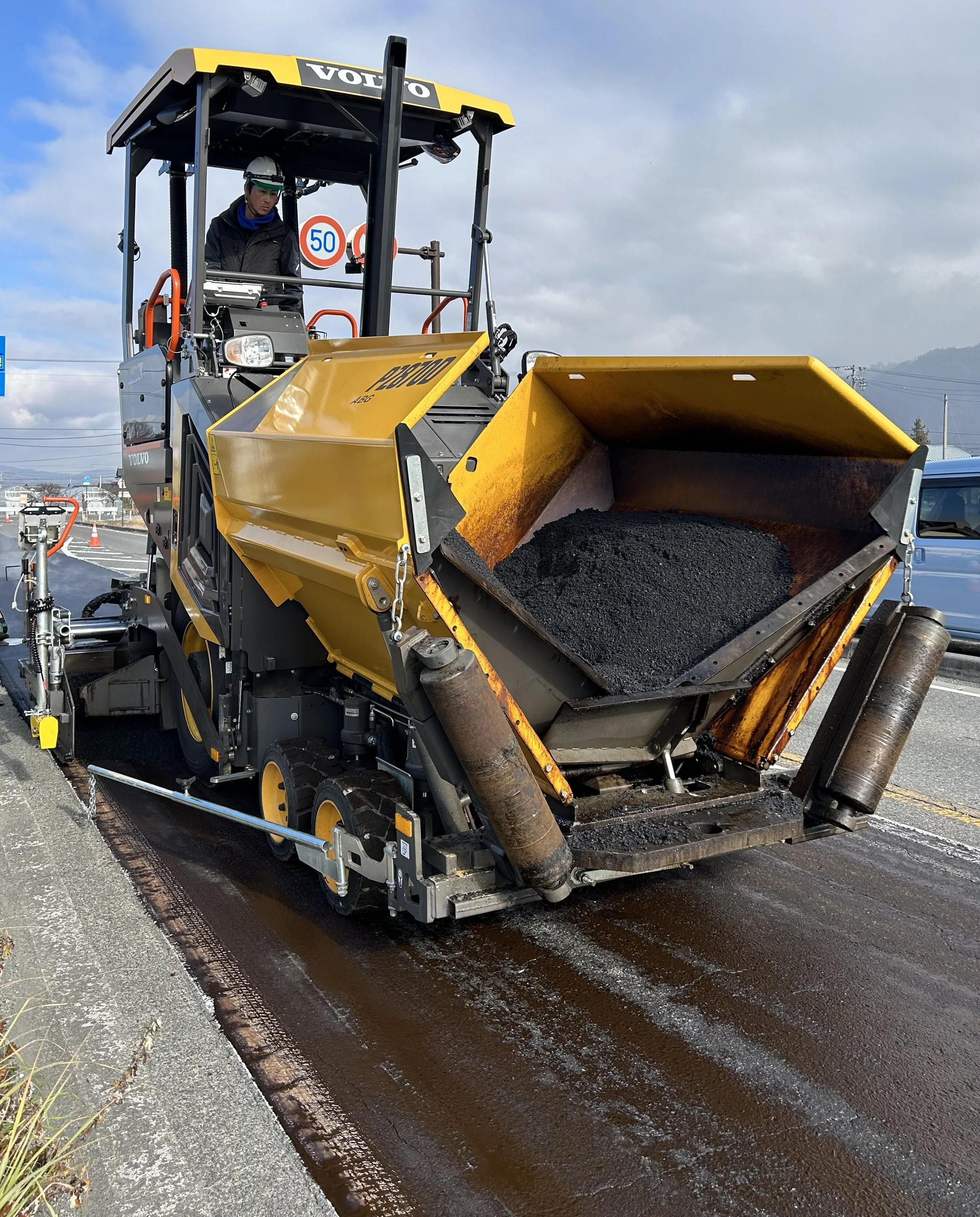 道路舗装にアスファルトを敷いている工事現場の写真。大型のダンプカーに似た機械がアスファルトを荷台に運び込んでいる。背景には山と空が見える。