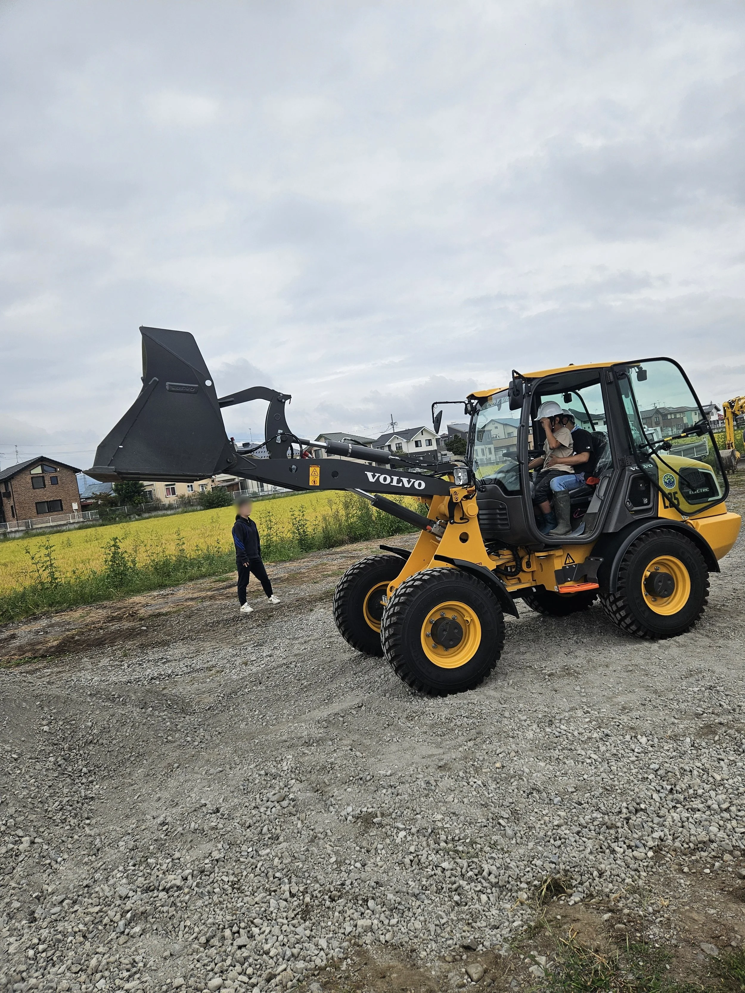 一信建設/黄色いボルボの小型建設機械が工事現場で働いている。操縦席には二人の作業員がおり、ヘルメットを着用している。背景には住宅の並ぶ景色と曇り空が見える。