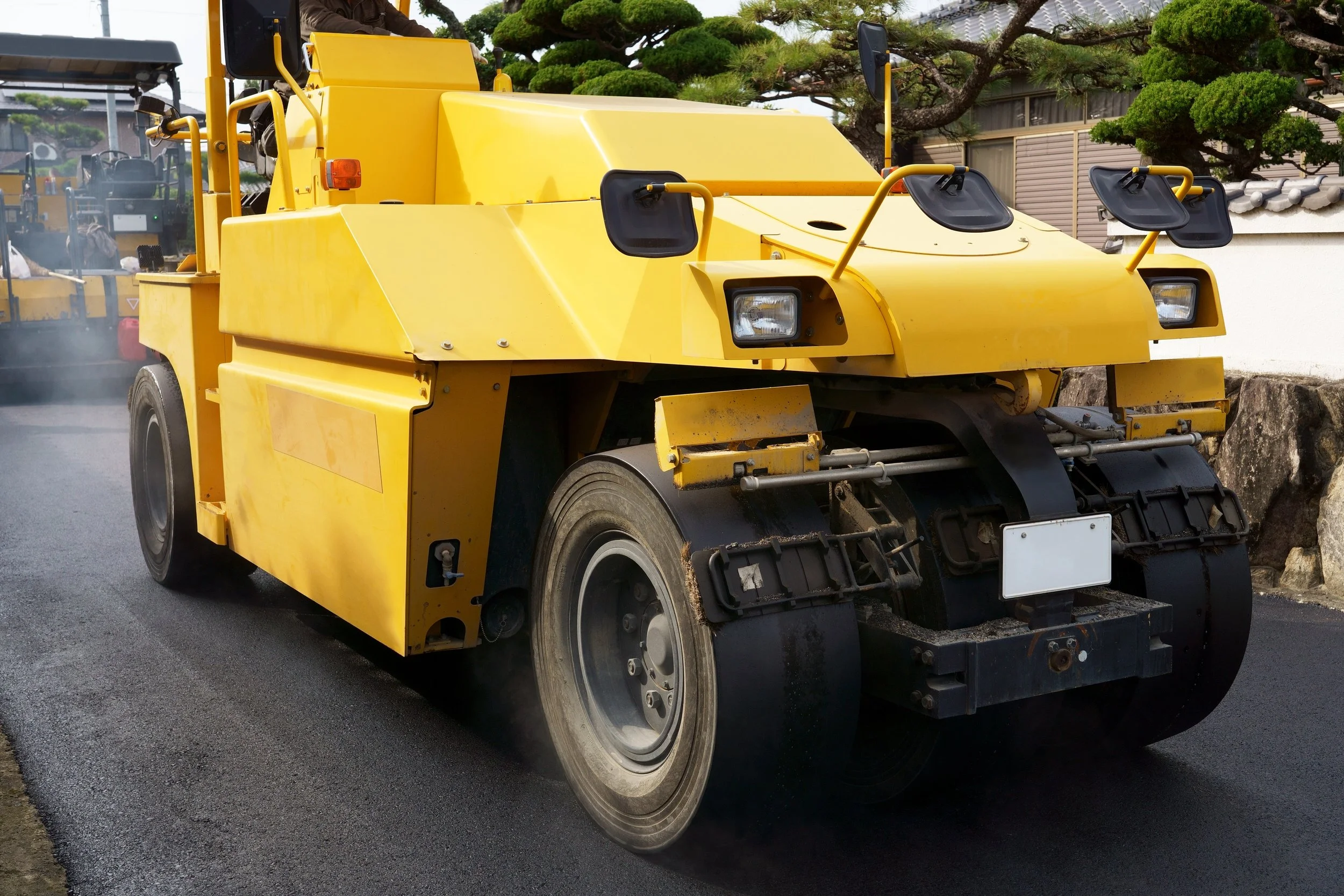 黄色いコンクリート舗装用の道路機械が道路を舗装している様子。周囲に緑の木や建物が見える。
