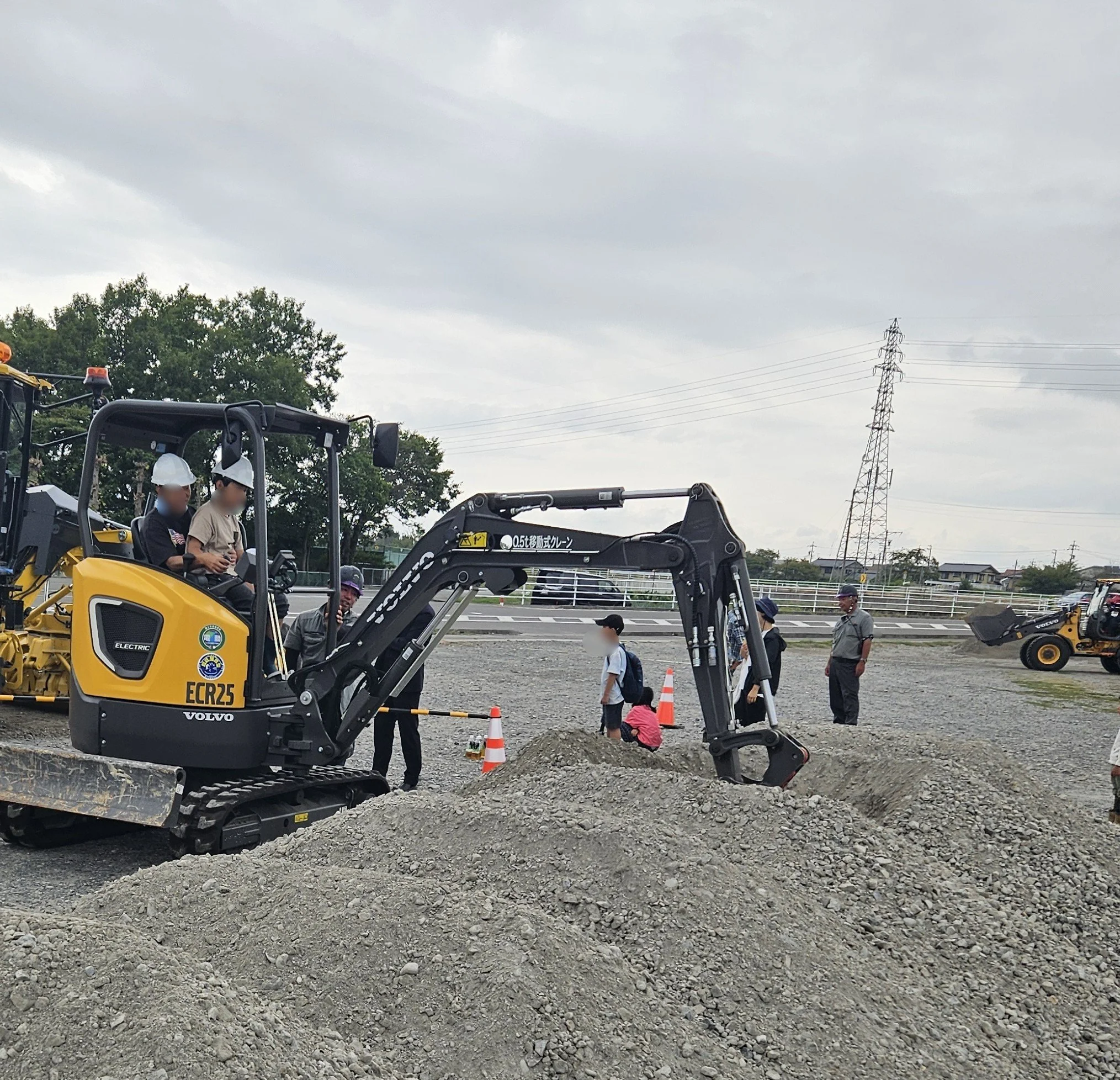一信建設/建設現場で働く人々と小型重機。空は曇り。背景には木と電柱が見える。