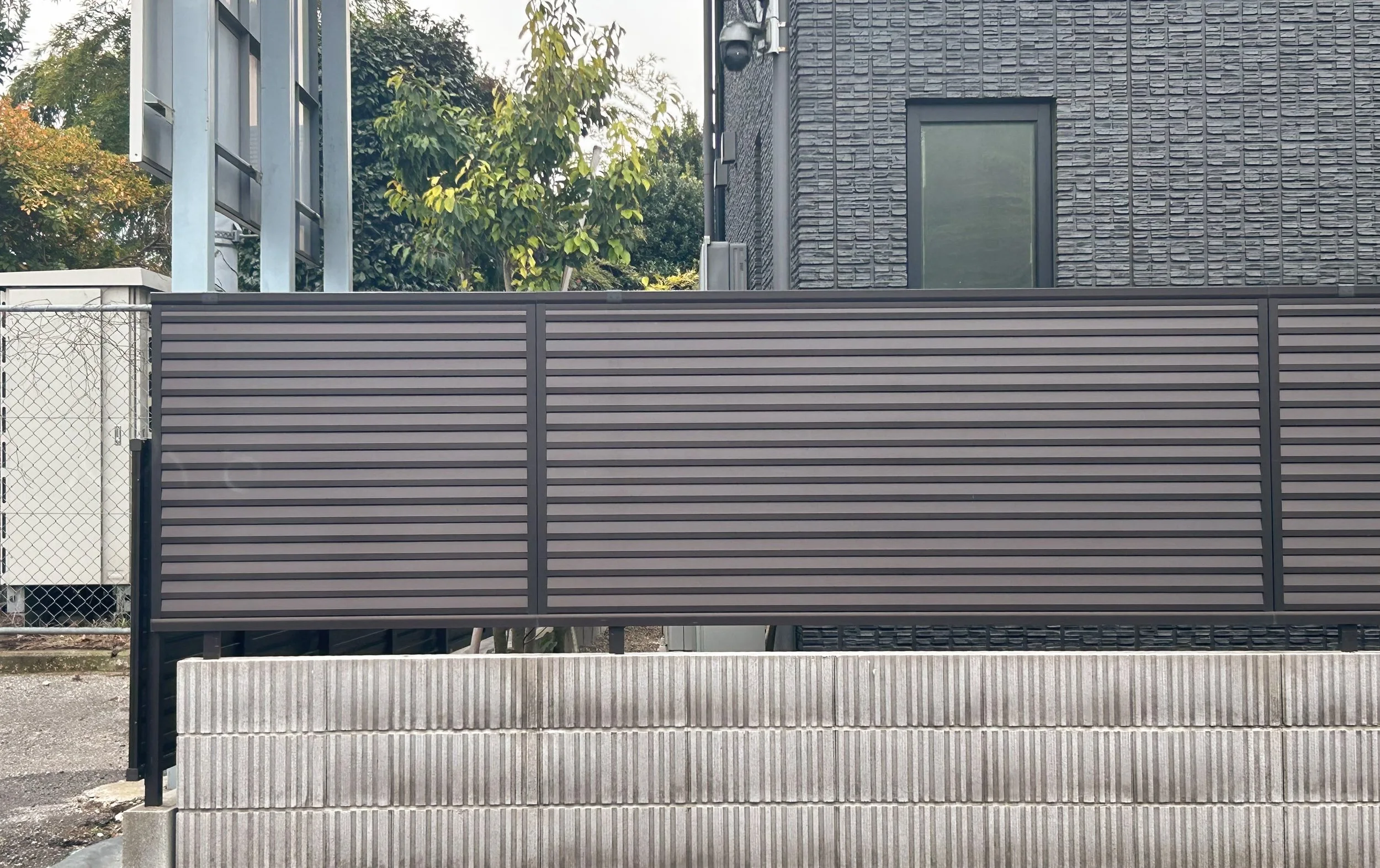 黒い壁の建物の側面とフェンスのある景色。左側に緑の葉を持つ木と一部の建物の屋根が見える。