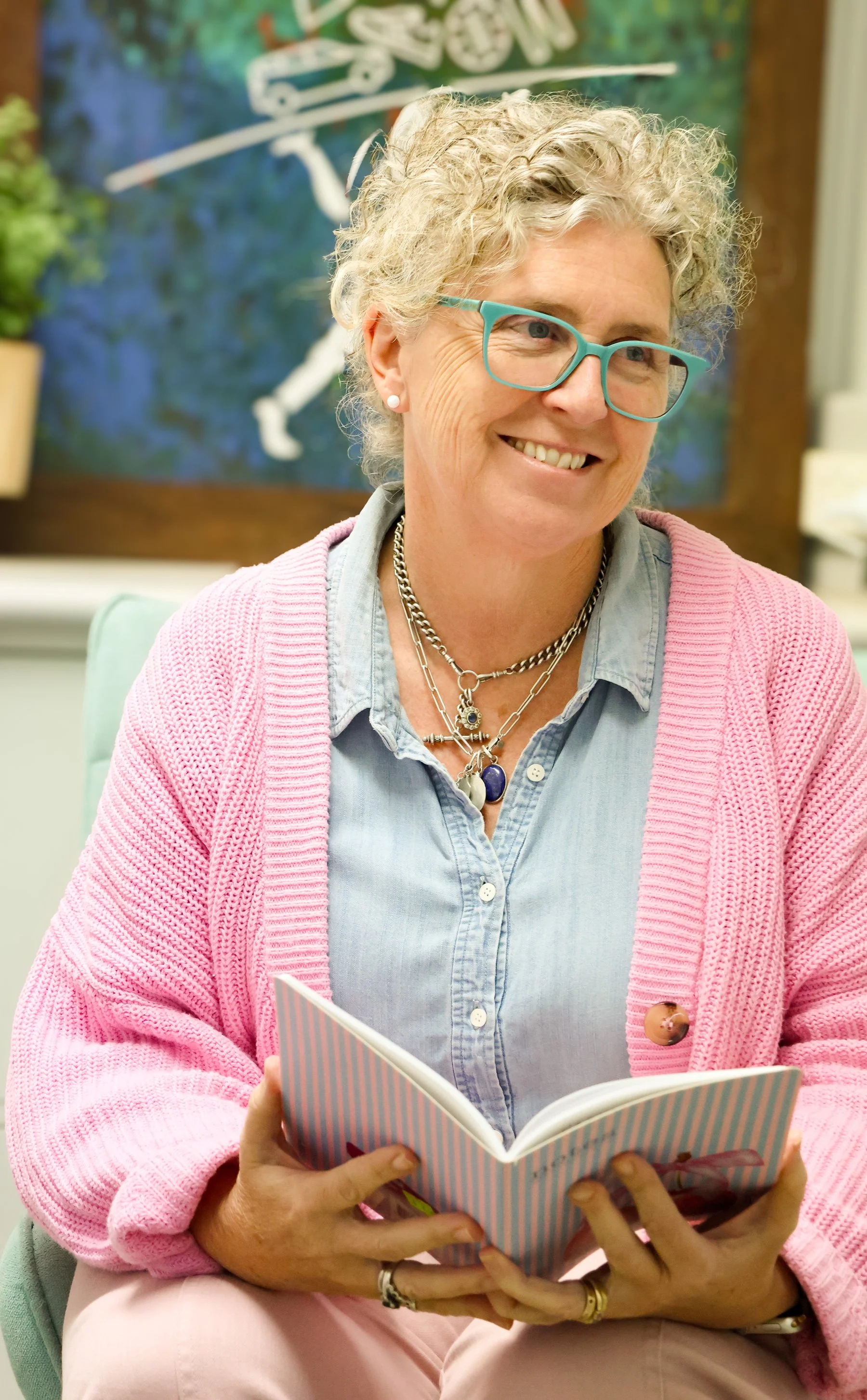 Fi Waters with curly gray hair and blue glasses, smiling while reading a book. She is wearing a denim shirt, a pink cardigan, multiple necklaces, and earrings. The background features a colorful abstract painting.