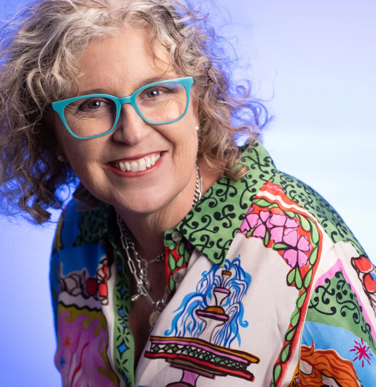 Fi Waters smiling, with curly grey hair, blue glasses, and a colorful, patterned shirt with floral and fountain designs.